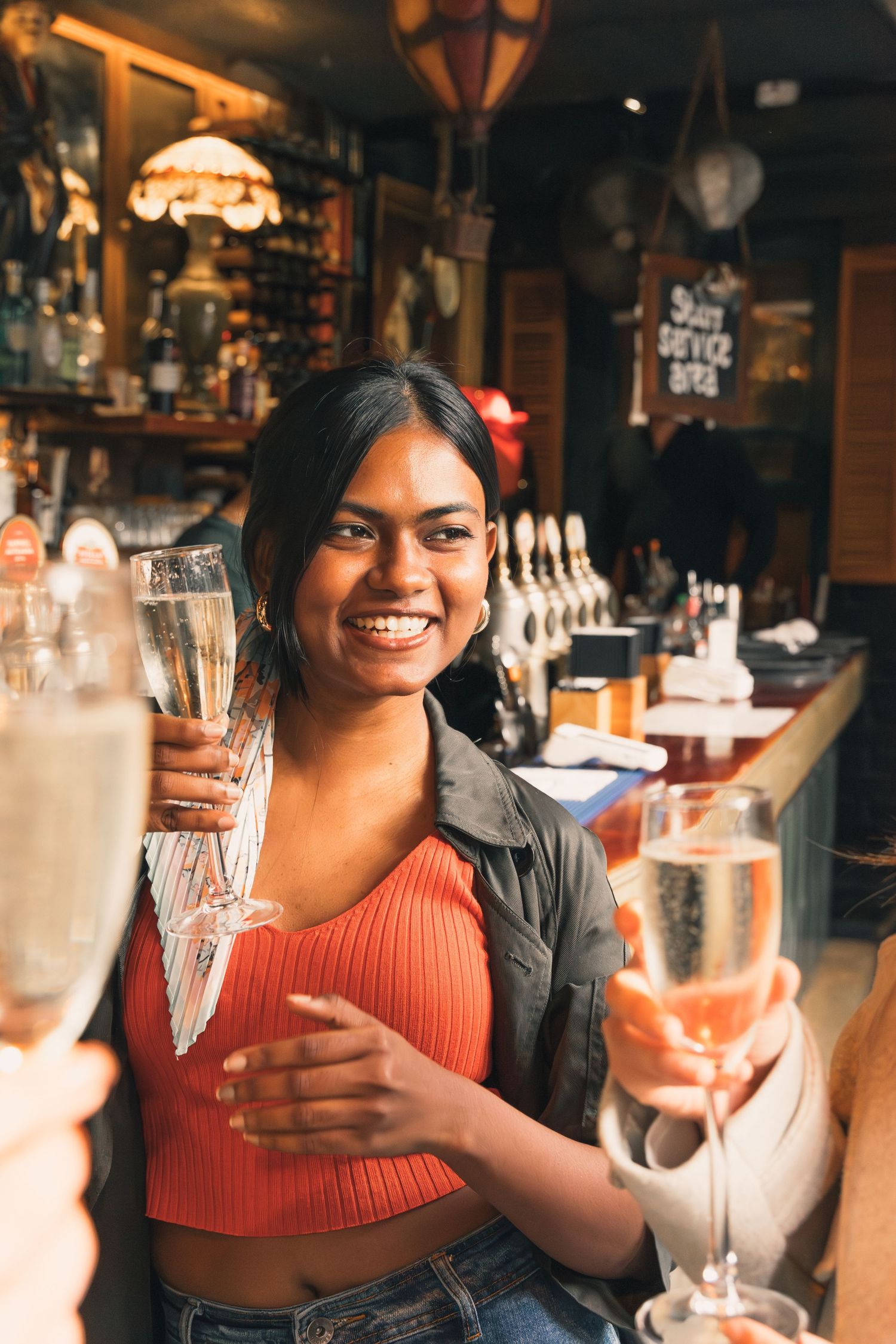Smiling young woman holding champagne flute at a bar, toasting with friends in a warmly lit pub.