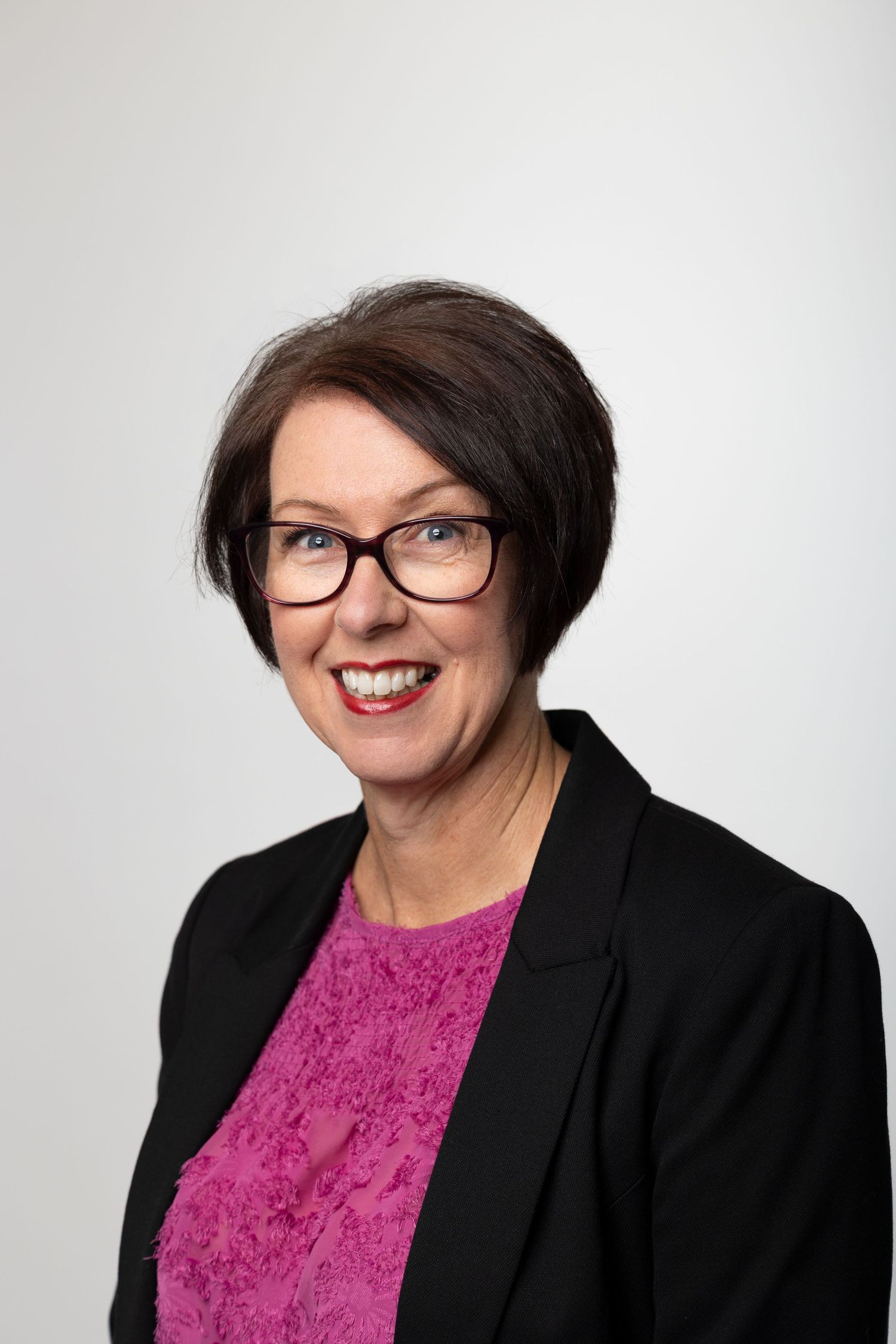 Professional headshot of a middle-aged woman with short dark hair and glasses wearing a pink top and black blazer.