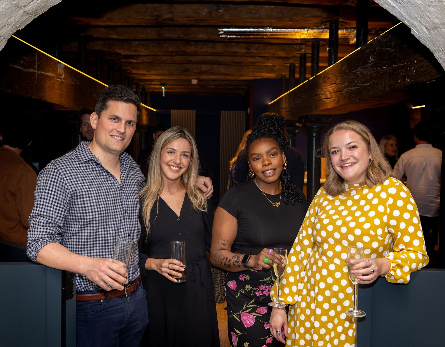 Four friends smile and hold drinks at an elegant indoor party event.