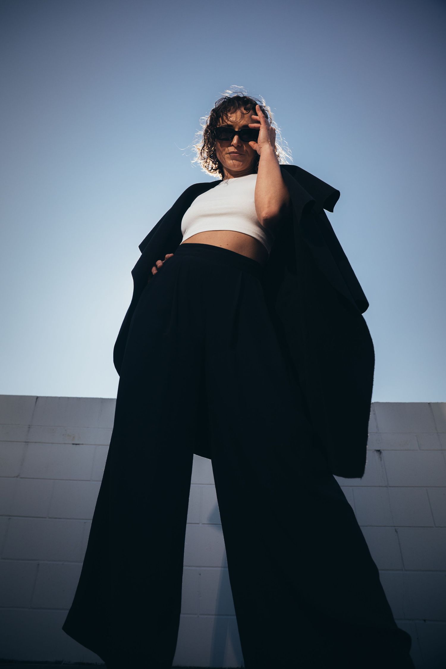 Stylish woman in black wide-leg pants and oversized blazer posing confidently against blue sky background.