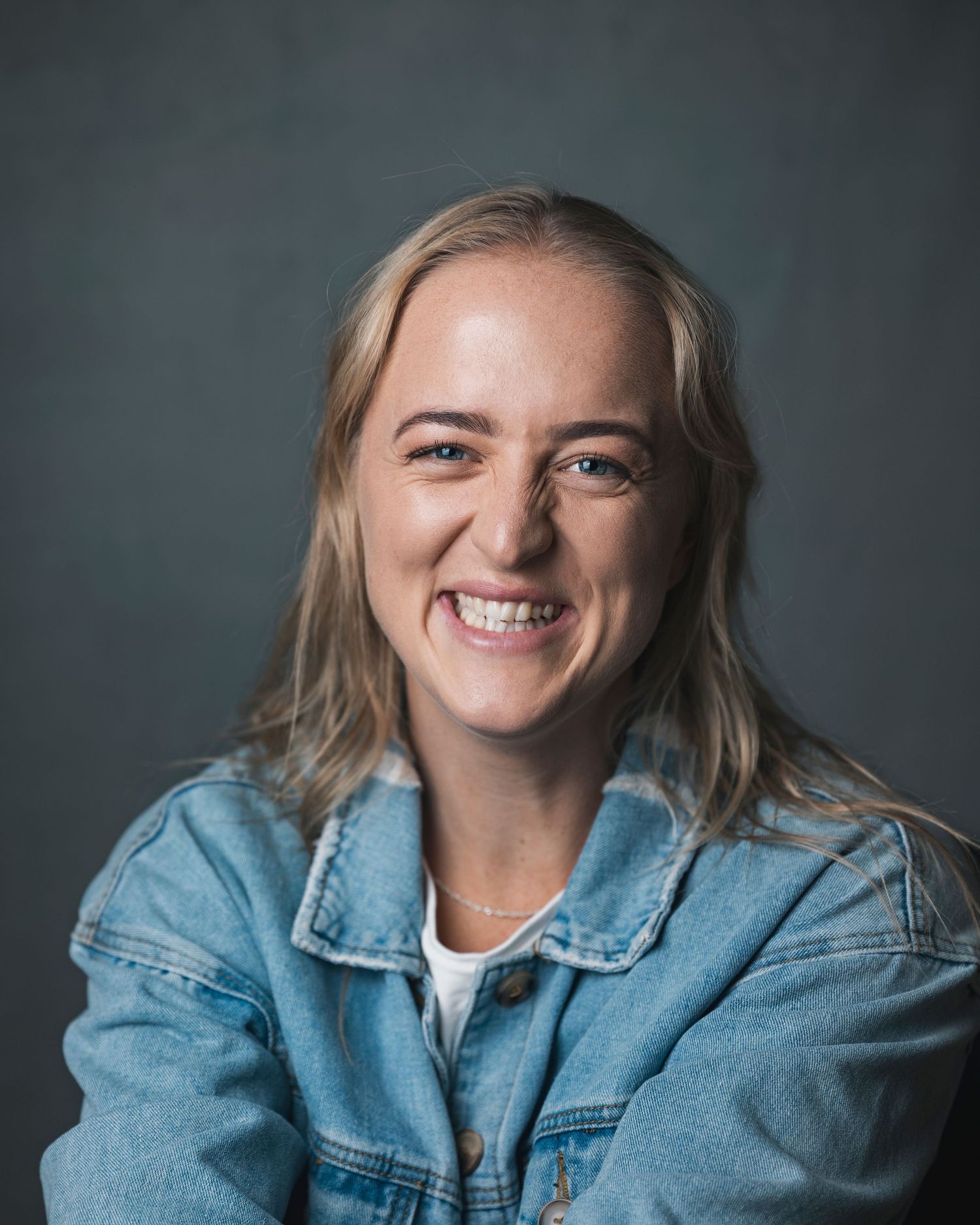 Smiling young woman with blonde hair wearing a denim jacket poses for a professional headshot against a gray background.
