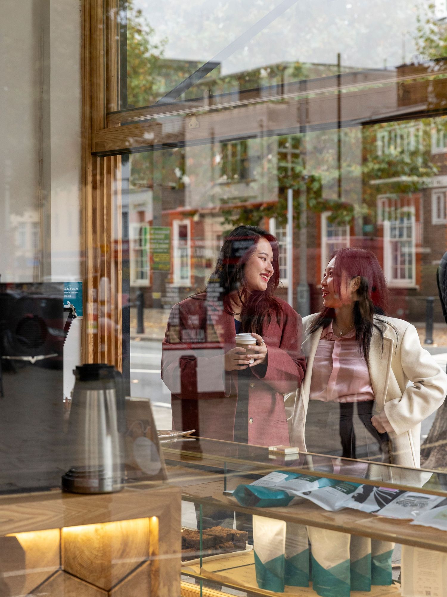 Two smiling women with coffee cups browsing inside a shop viewed through a large storefront window.