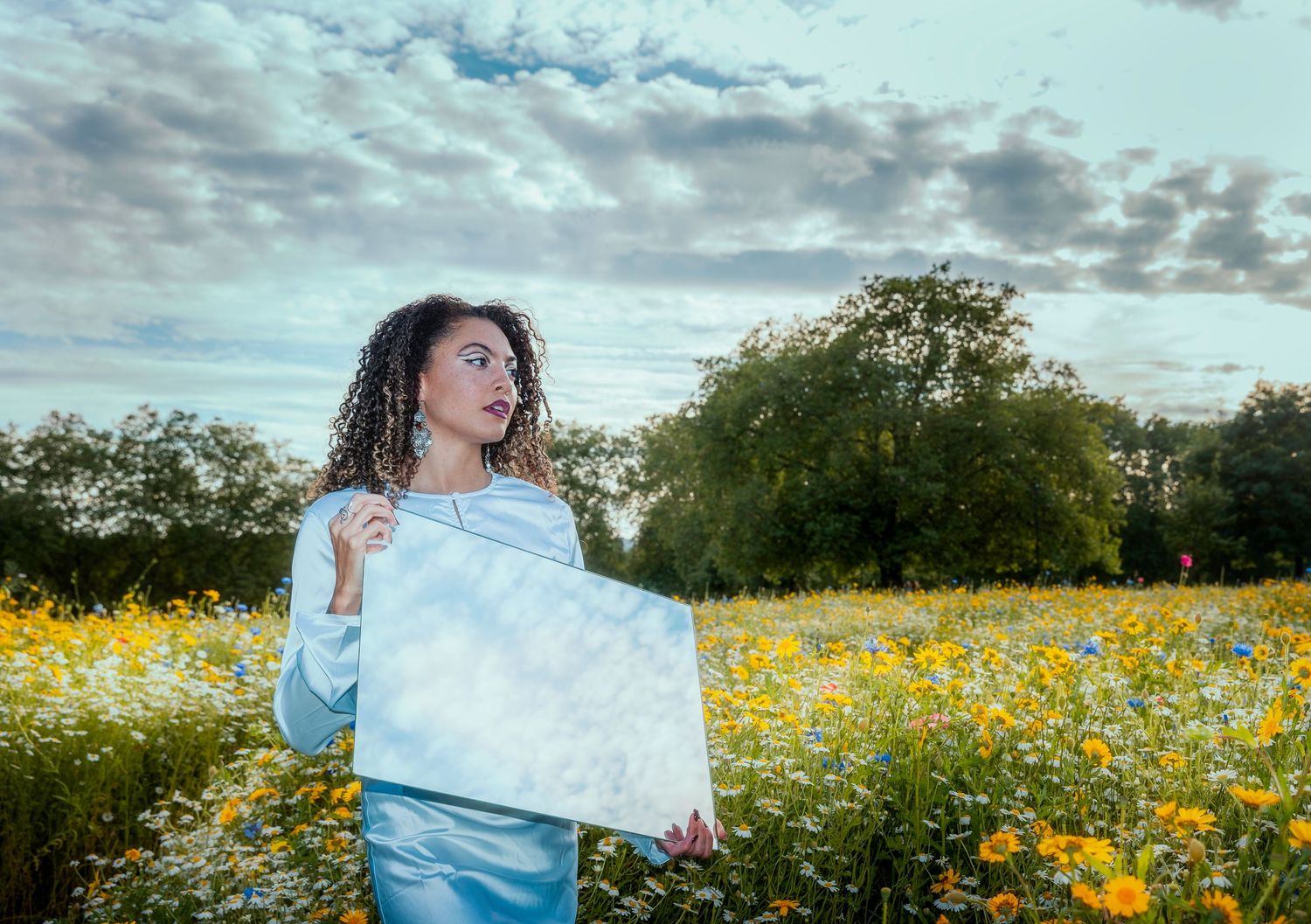 Woman in light blue outfit holding canvas in vibrant wildflower meadow under cloudy sky.