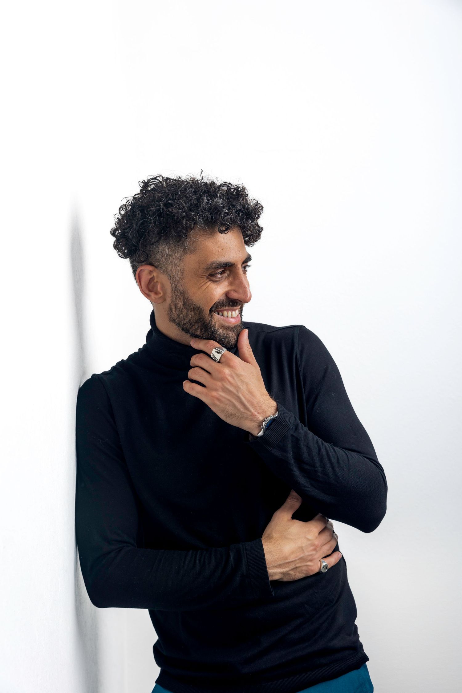 Smiling bearded man with curly hair in black turtleneck leaning against white wall.