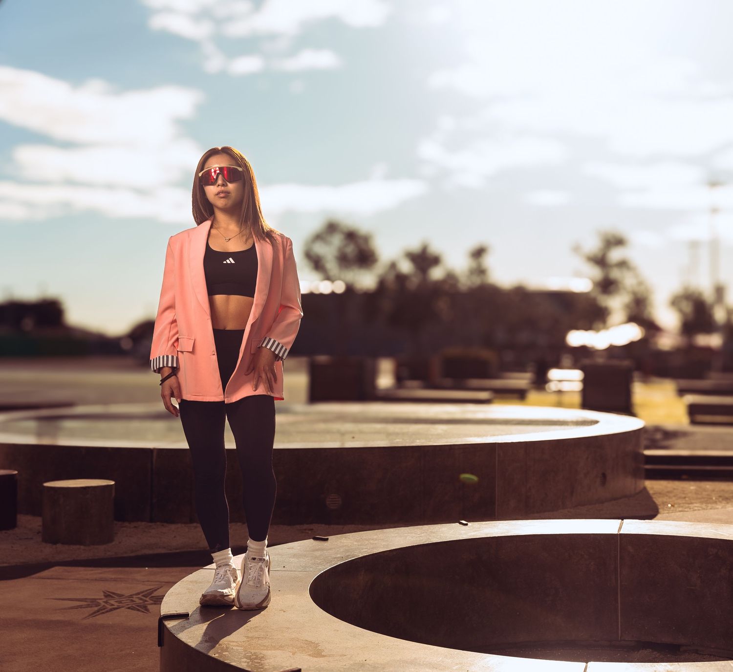 Stylish woman in pink blazer and black leggings standing near a circular fountain outdoors.
