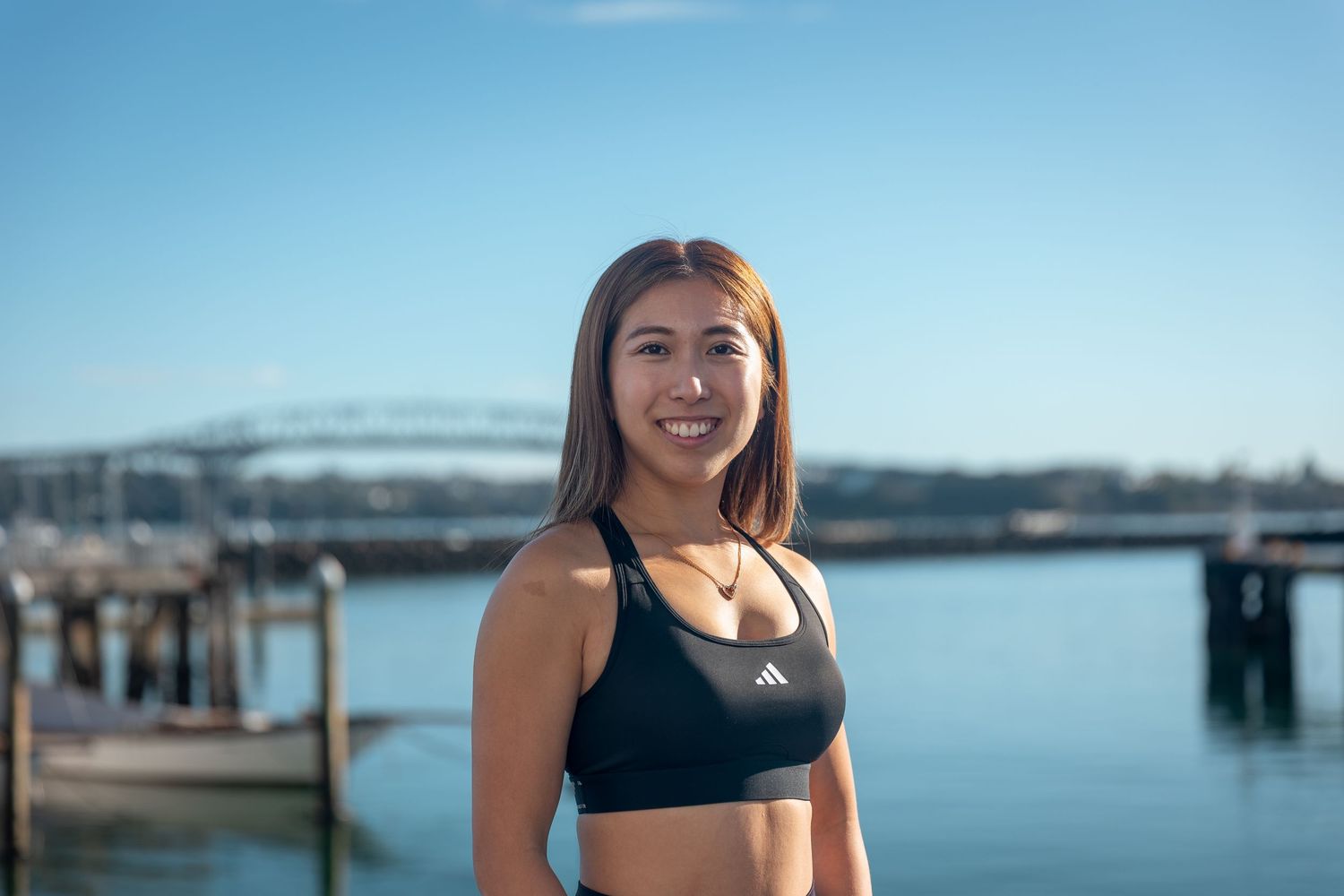 Smiling Asian woman in black Adidas sports bra standing in front of a harbor bridge on a sunny day.