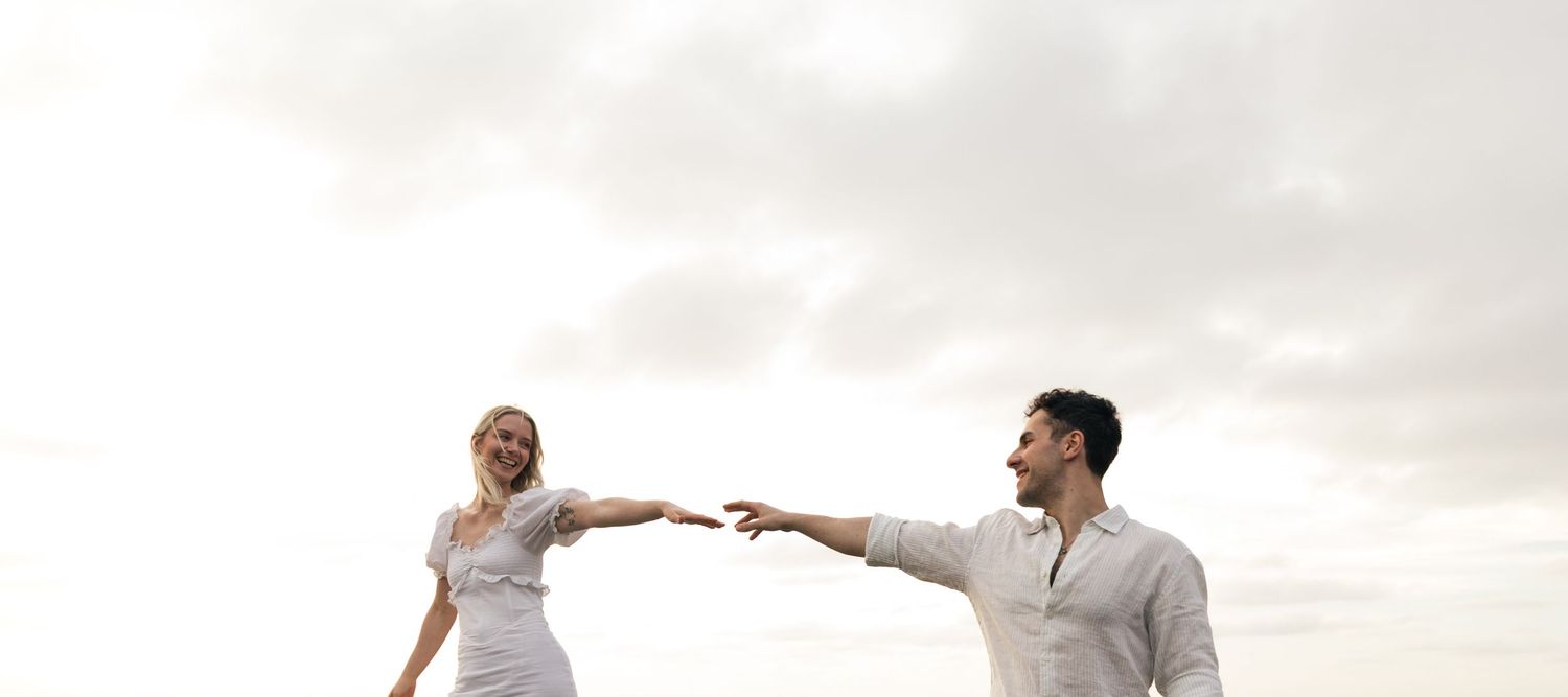 Couple reaching toward each other against cloudy sky, woman in white dress laughing joyfully outdoors.