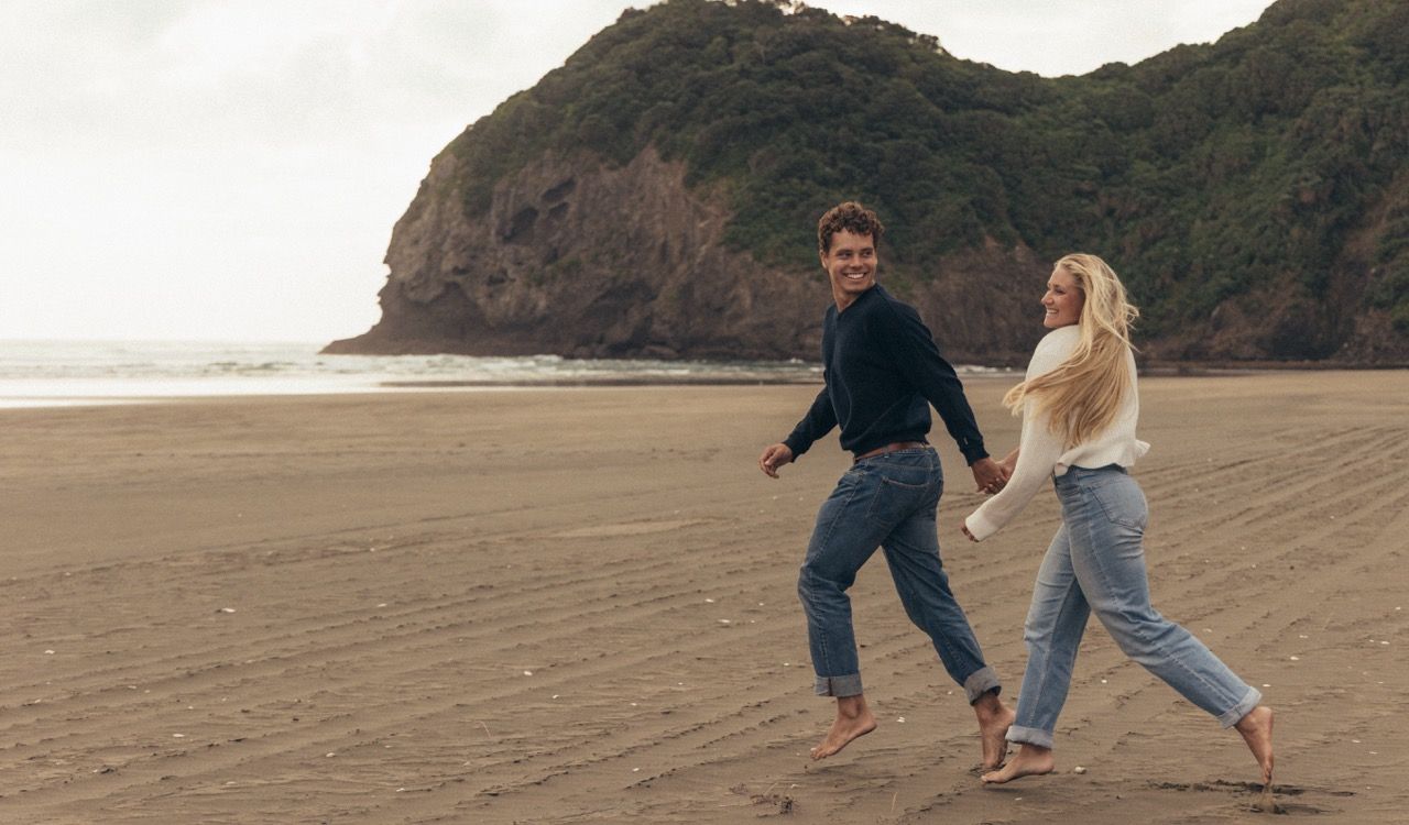 Couple holding hands and walking barefoot on a dark sandy beach with a rocky headland in the background.