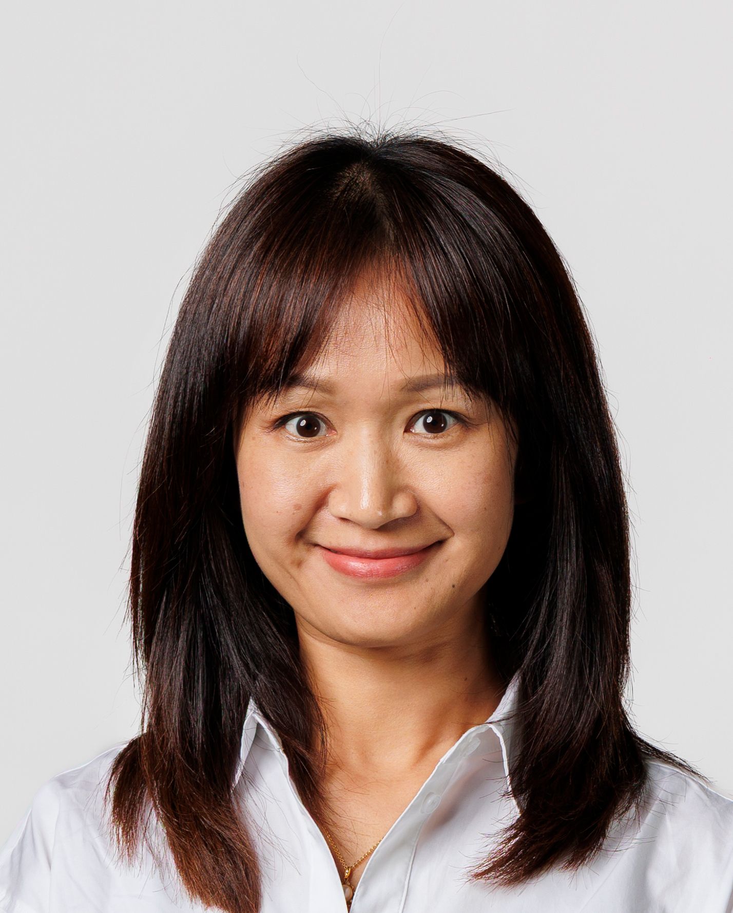 Smiling woman with dark brown hair and bangs wearing a white collared shirt against a gray background.