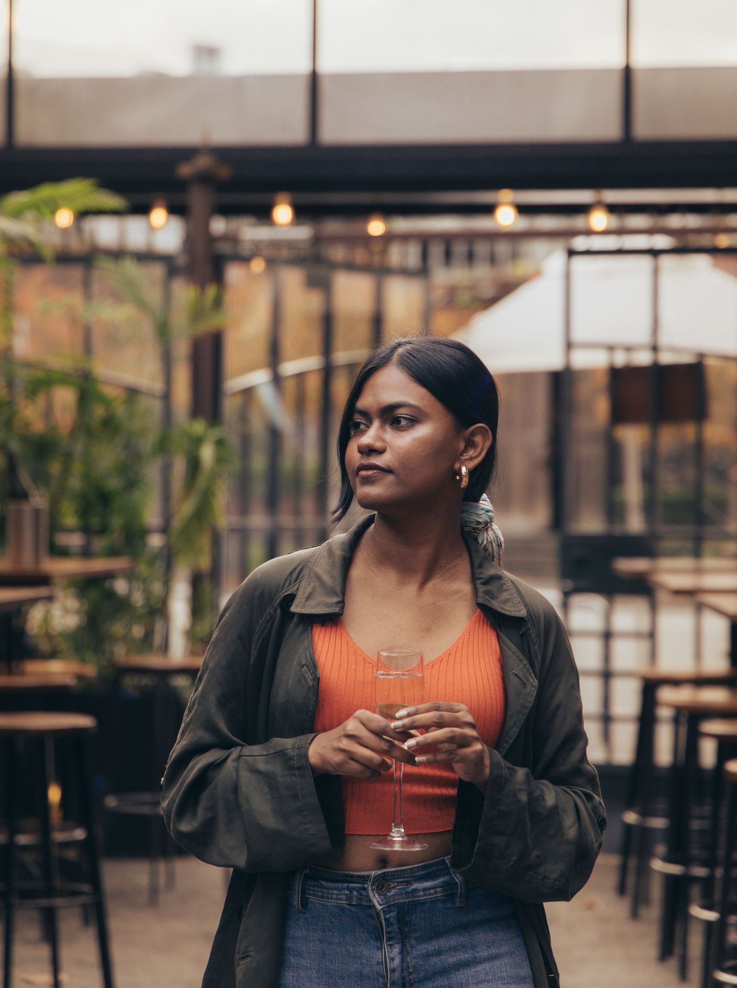 Young woman in orange crop top and olive jacket holds a cocktail at a stylish bar patio.