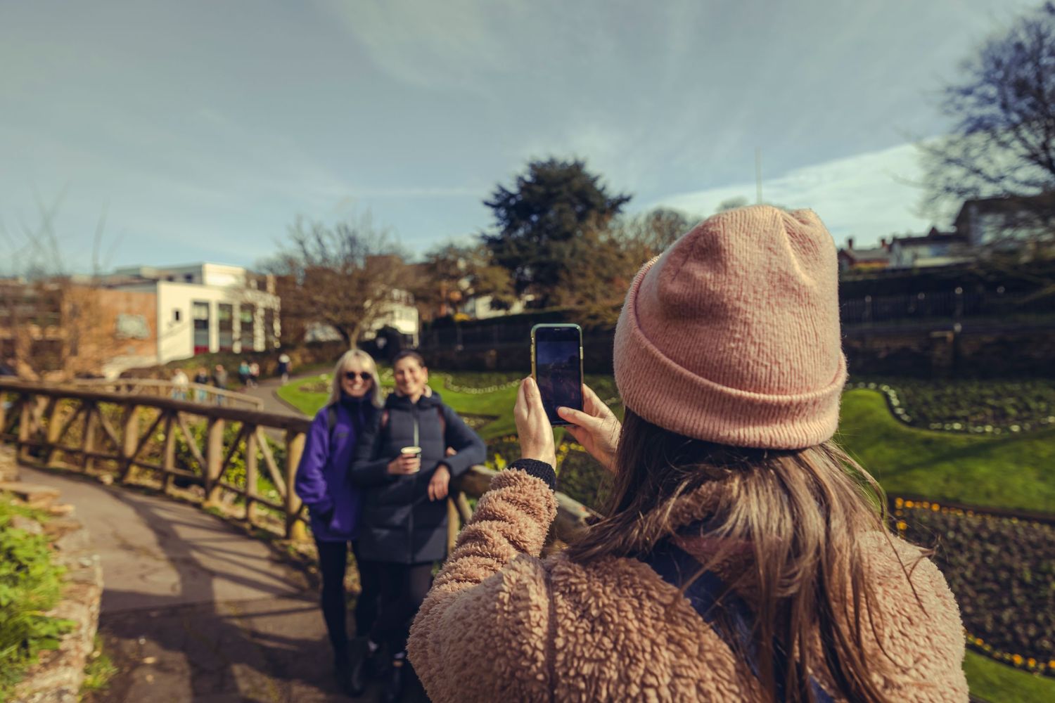 Woman in pink hat photographing two friends on a bridge in a park.