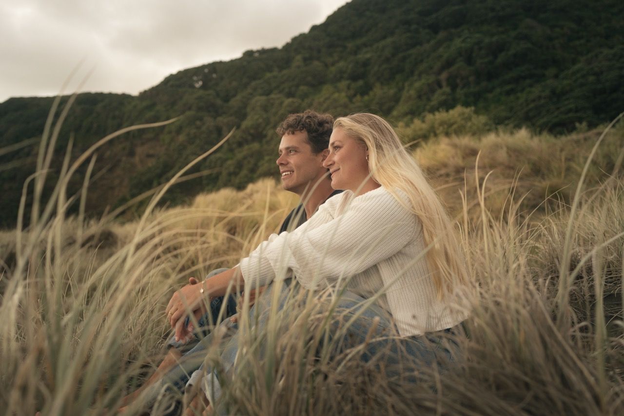 Couple sitting together in tall grass dunes with lush green hills in the background on a cloudy day.