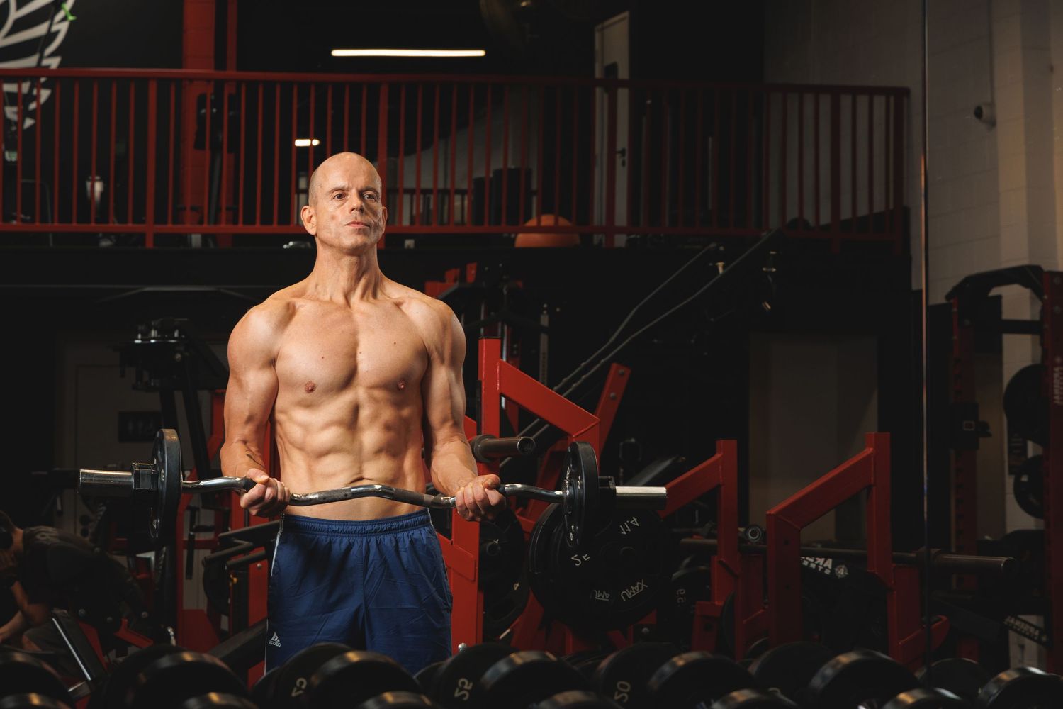 Muscular bald man lifting barbell in gym, showcasing defined abs and athletic physique during intense weightlifting workout.