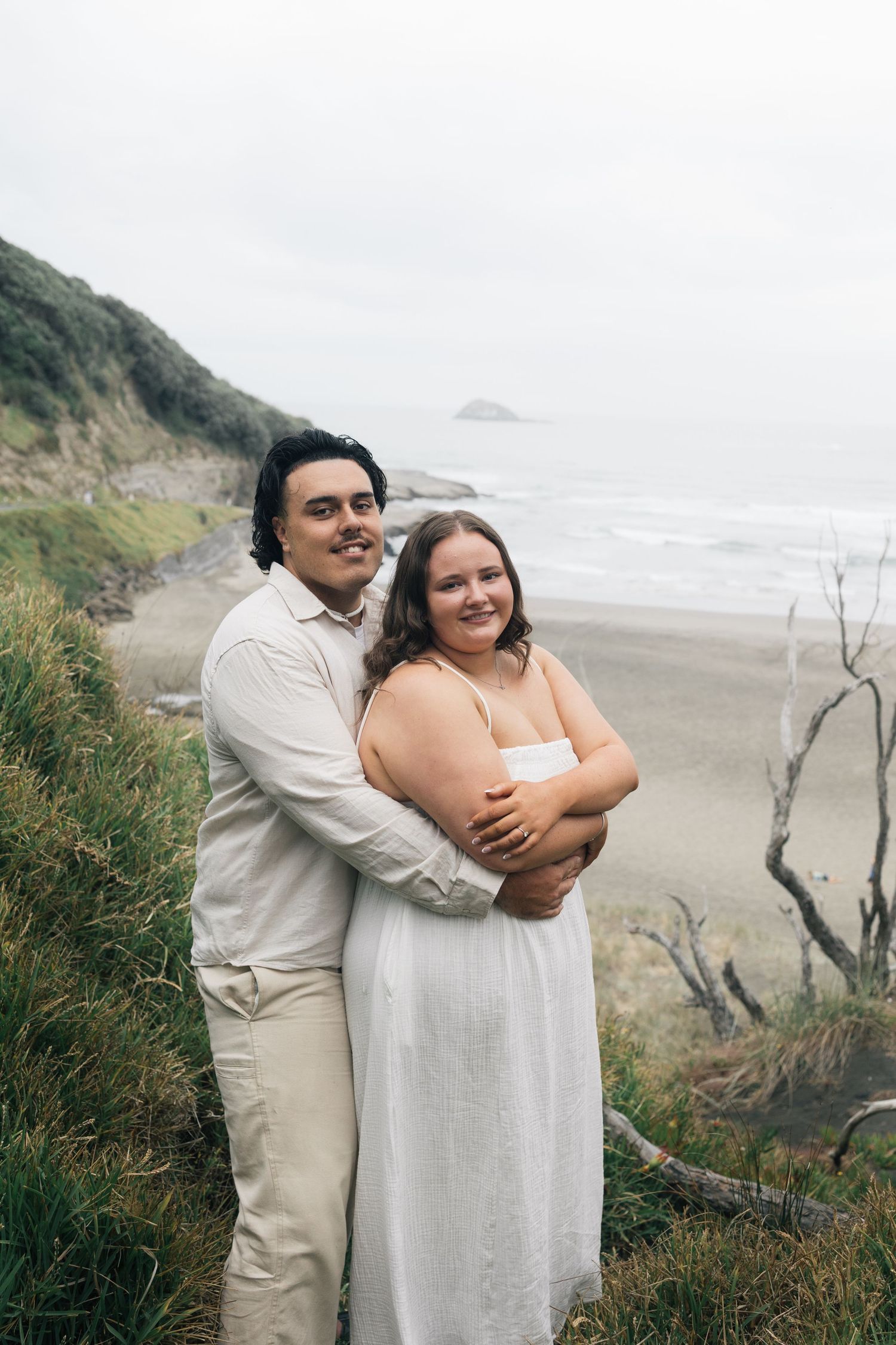 Smiling couple embracing on a grassy coastal cliff with a sandy beach backdrop.