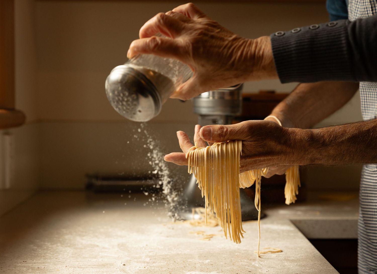 Hands dusting fresh homemade pasta with flour from a shaker over a floured wooden surface.