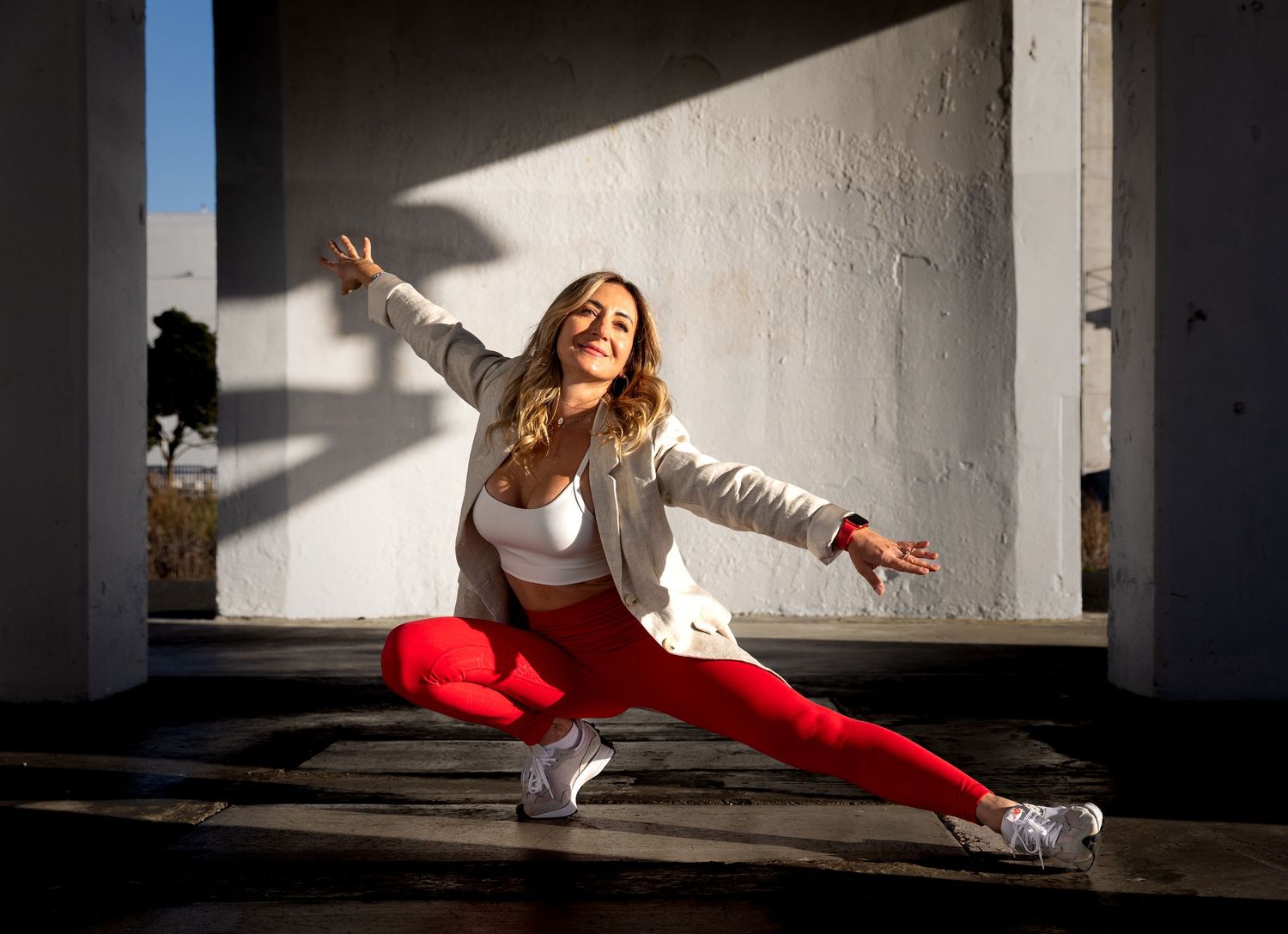 Blonde woman in red leggings doing a wide split pose in sunny urban setting.