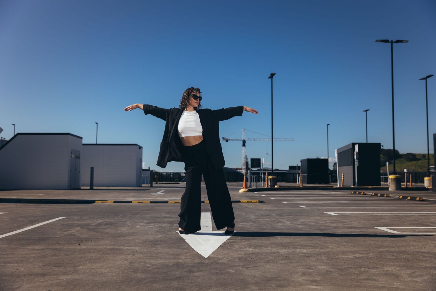 Woman in black blazer and white crop top posing with arms outstretched in sunny outdoor parking lot.