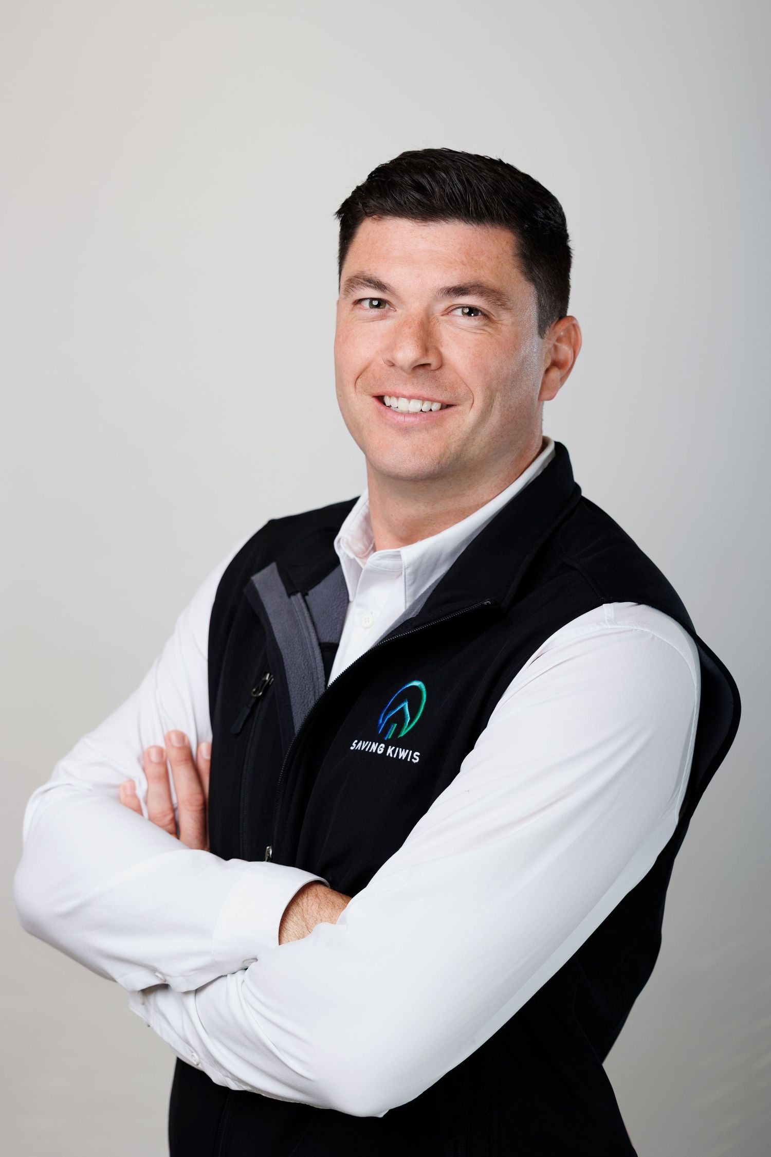 A smiling man in a Saving Kiwis vest stands with arms crossed against a light grey background.
