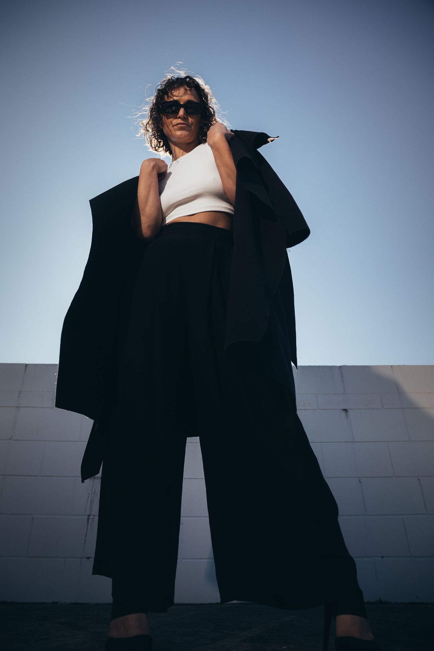 Woman in sunglasses wearing black wide-leg pants and oversized coat poses against white brick wall in sunlight.