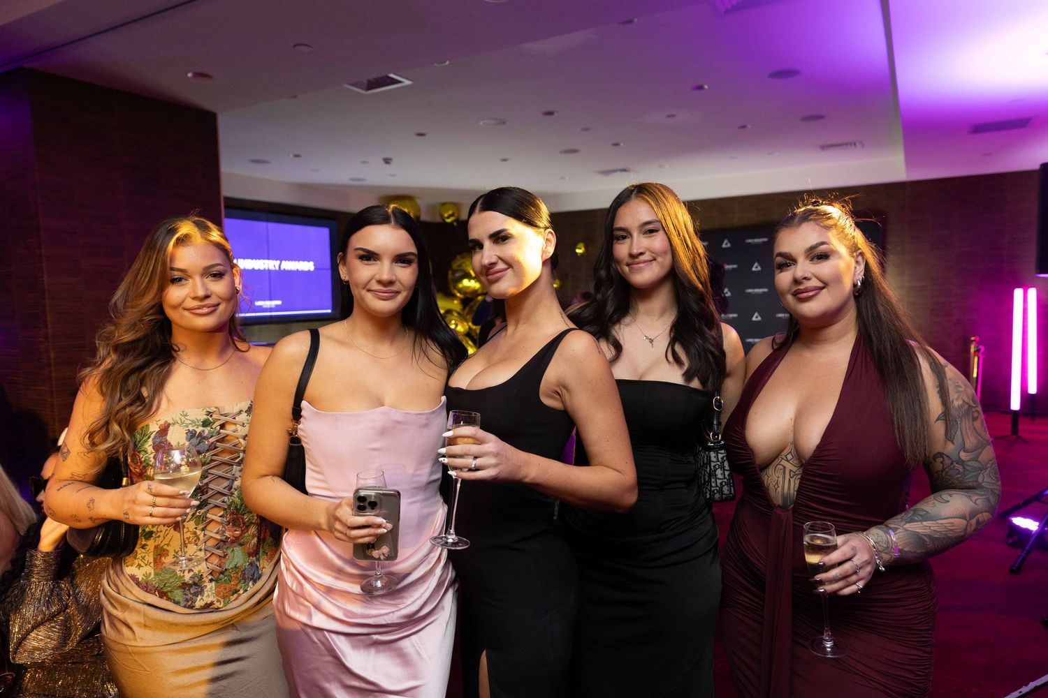 Five glamorously dressed women pose together holding champagne glasses at an upscale evening event.