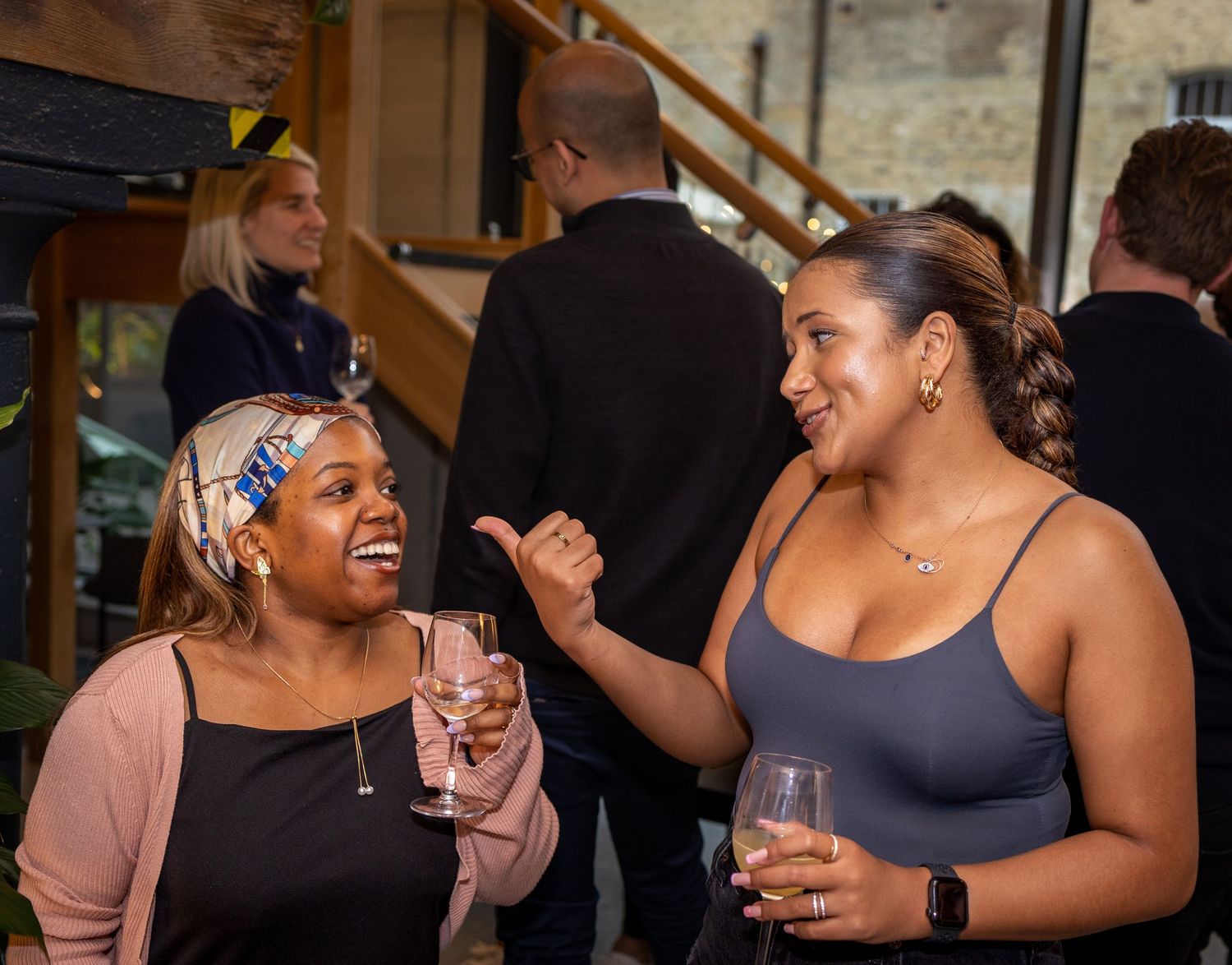 Two women laugh and chat while holding pink drinks at a lively indoor social event.
