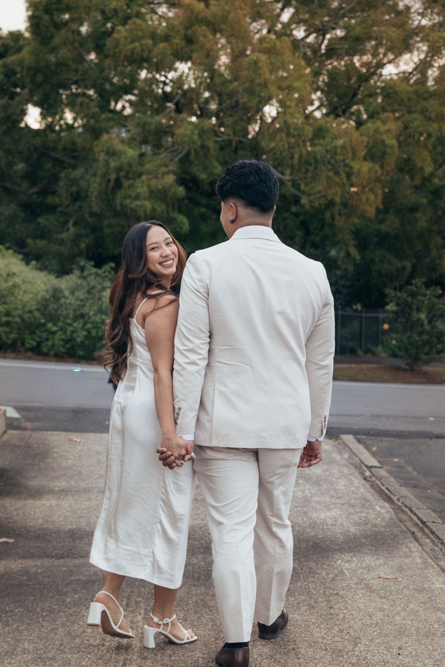 Couple in matching white outfits holding hands outdoors, woman smiling back at camera, lush green trees in background.