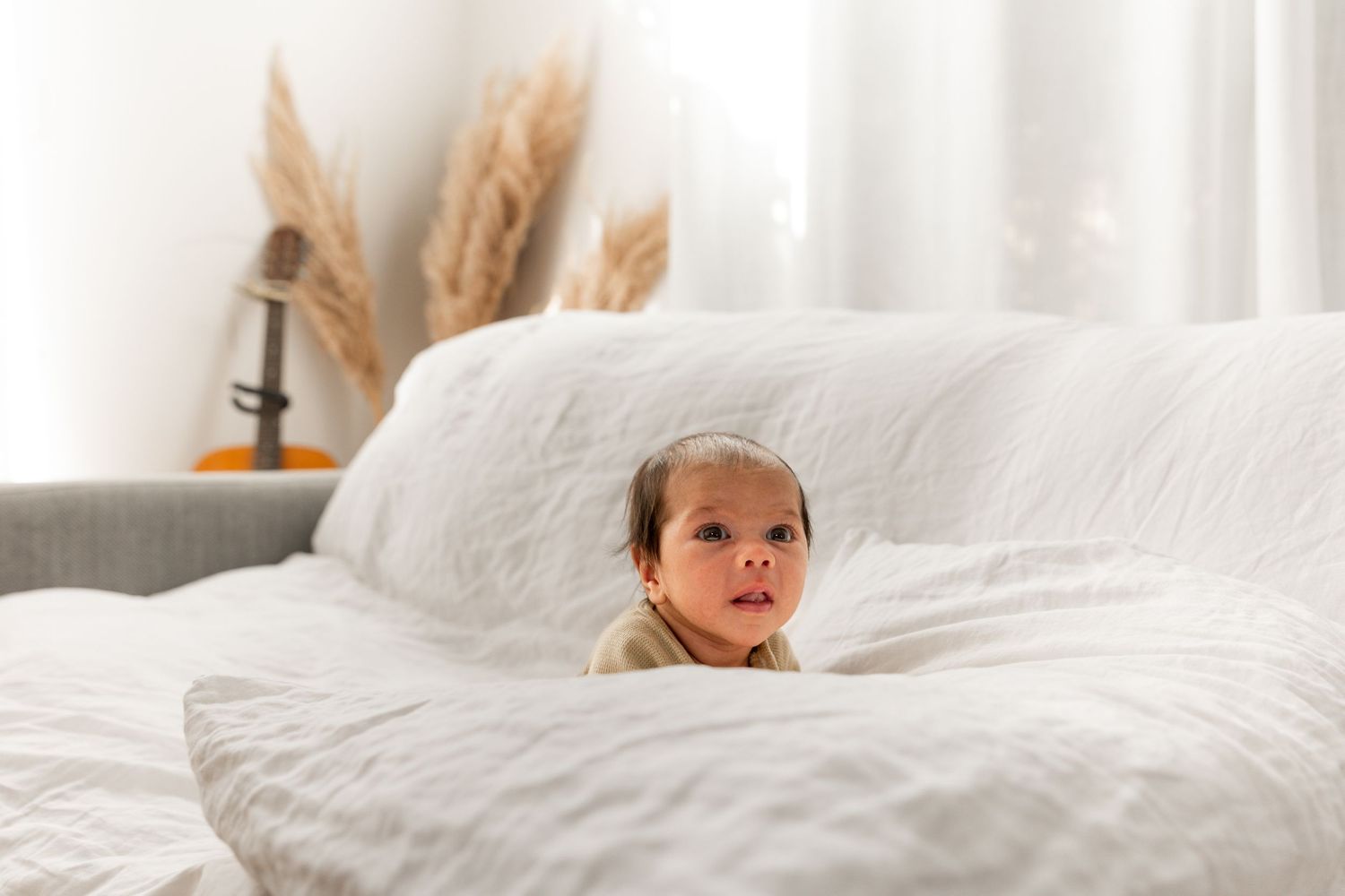 A curious baby peeks over a white fluffy bed with pampas grass decor in a bright, airy room.