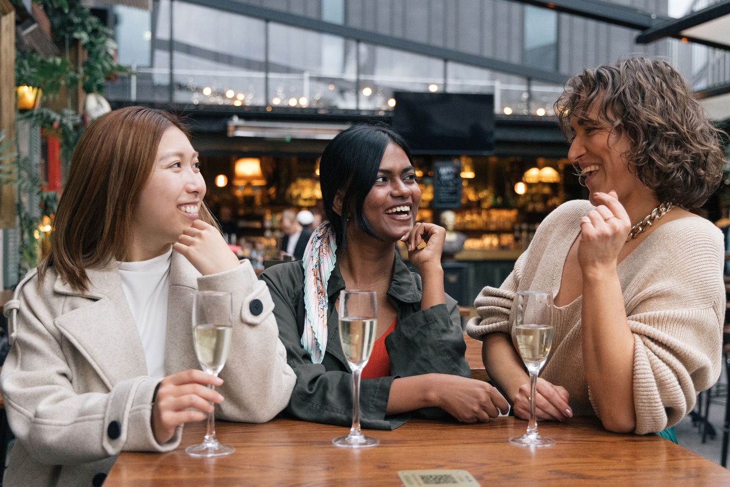 Three women laughing and enjoying champagne together at a wooden table in a lively bar.