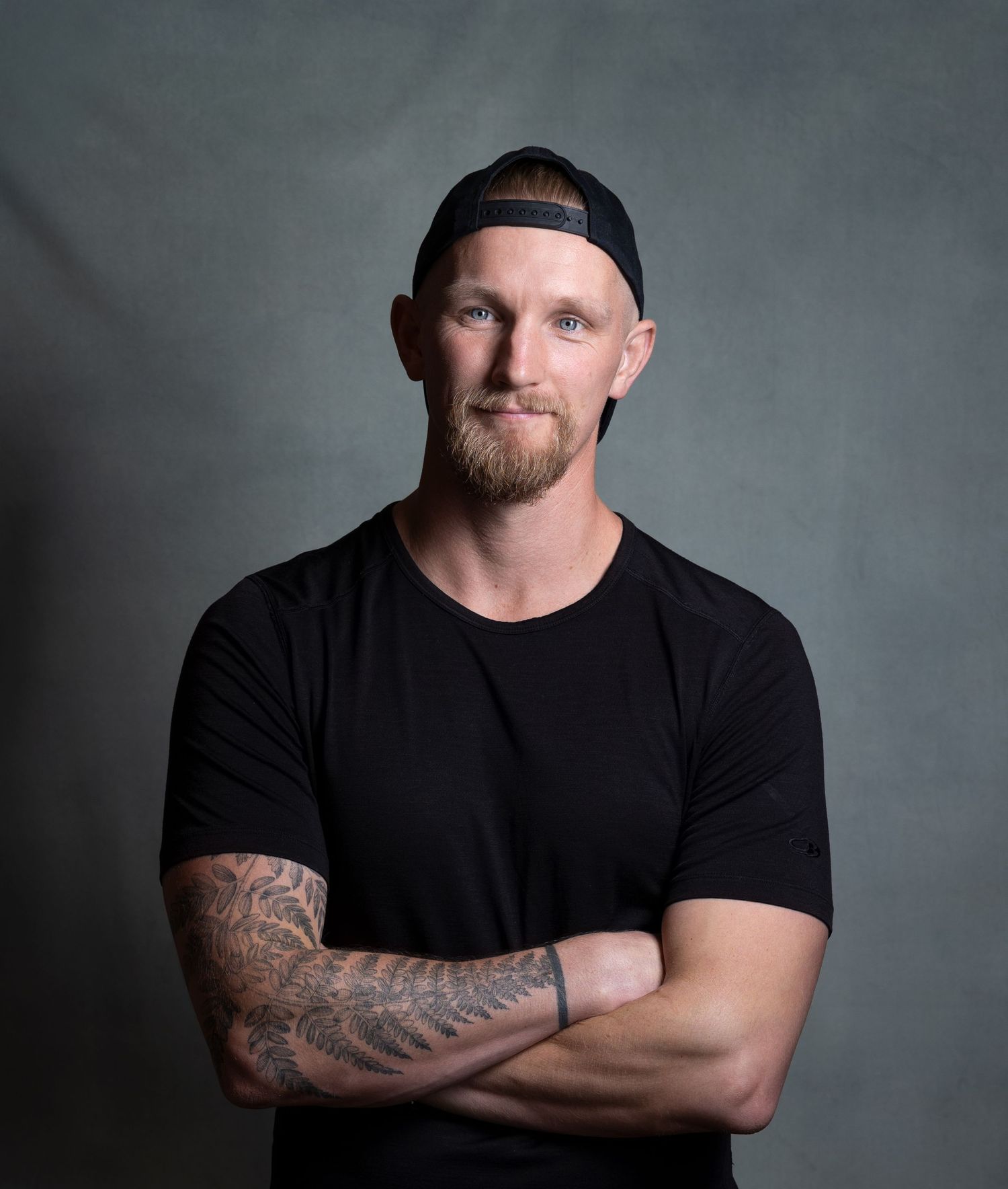 Muscular tattooed man in black t-shirt with arms crossed wearing backwards cap.