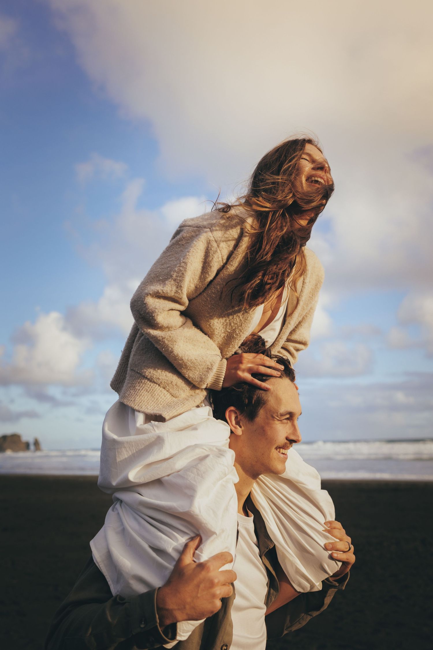 Woman sitting on mans shoulders laughing on a beach with dramatic cloudy sky and ocean backdrop.