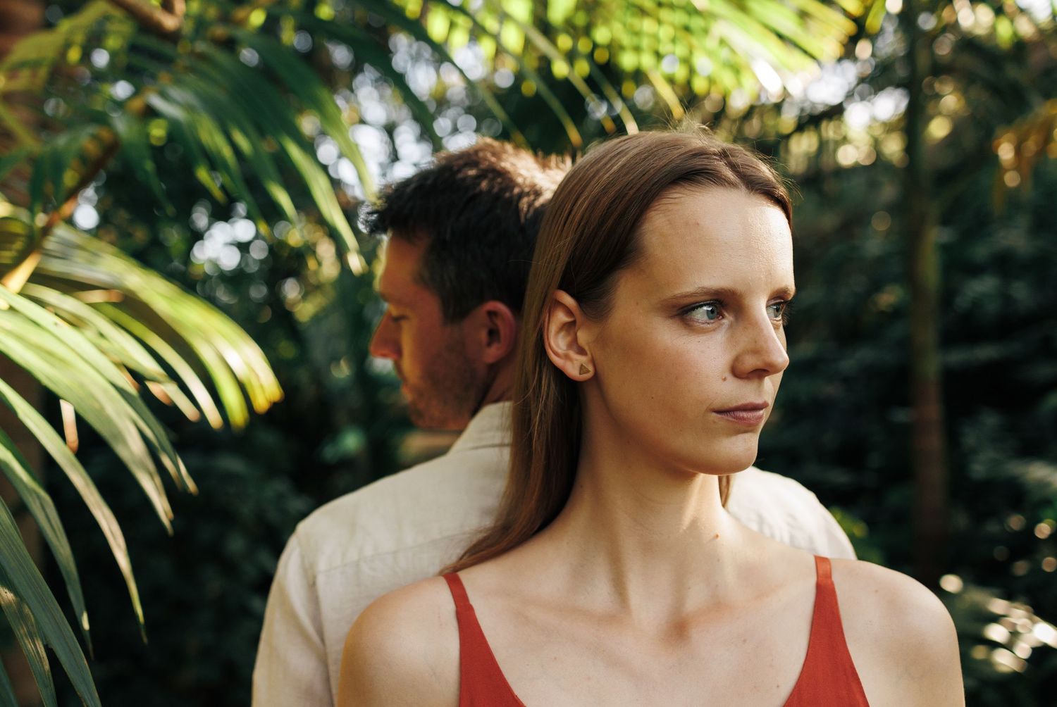 Young woman with straight hair gazing forward in tropical garden setting with man standing behind her.