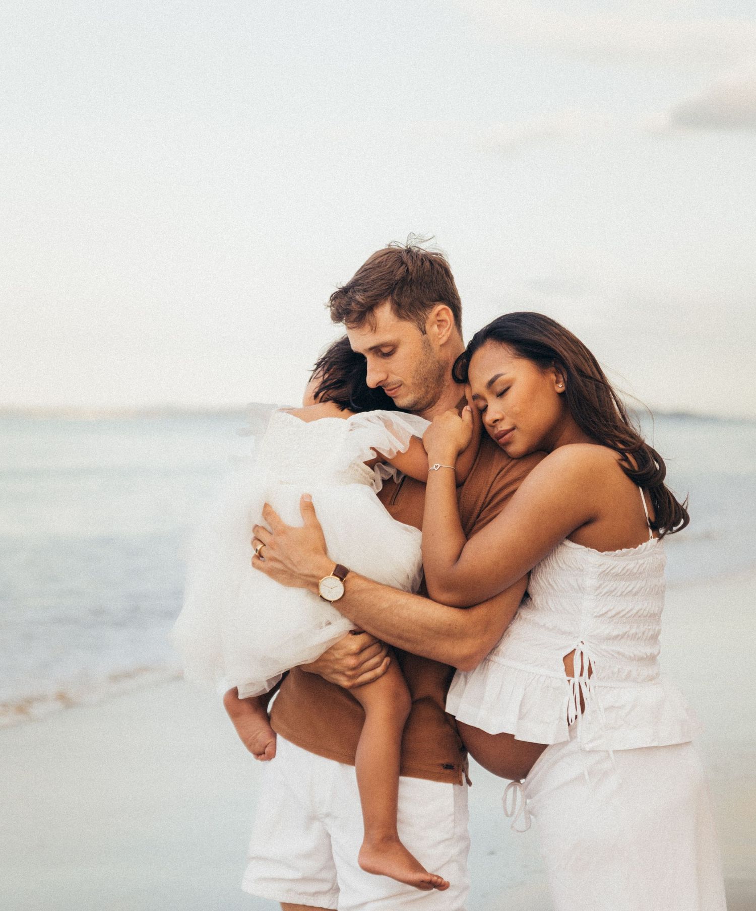 A loving family of three embraces on a tropical beach, dressed in white outfits, sharing a tender moment.