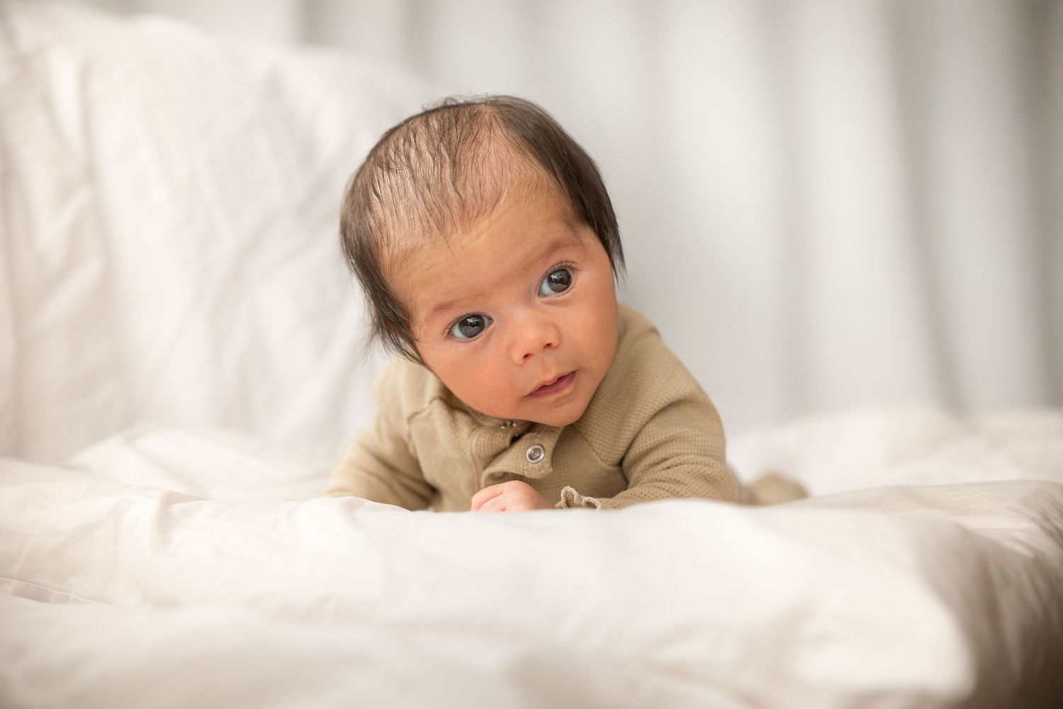 A newborn baby in an olive onesie doing tummy time on a white fluffy blanket, looking curiously at the camera.