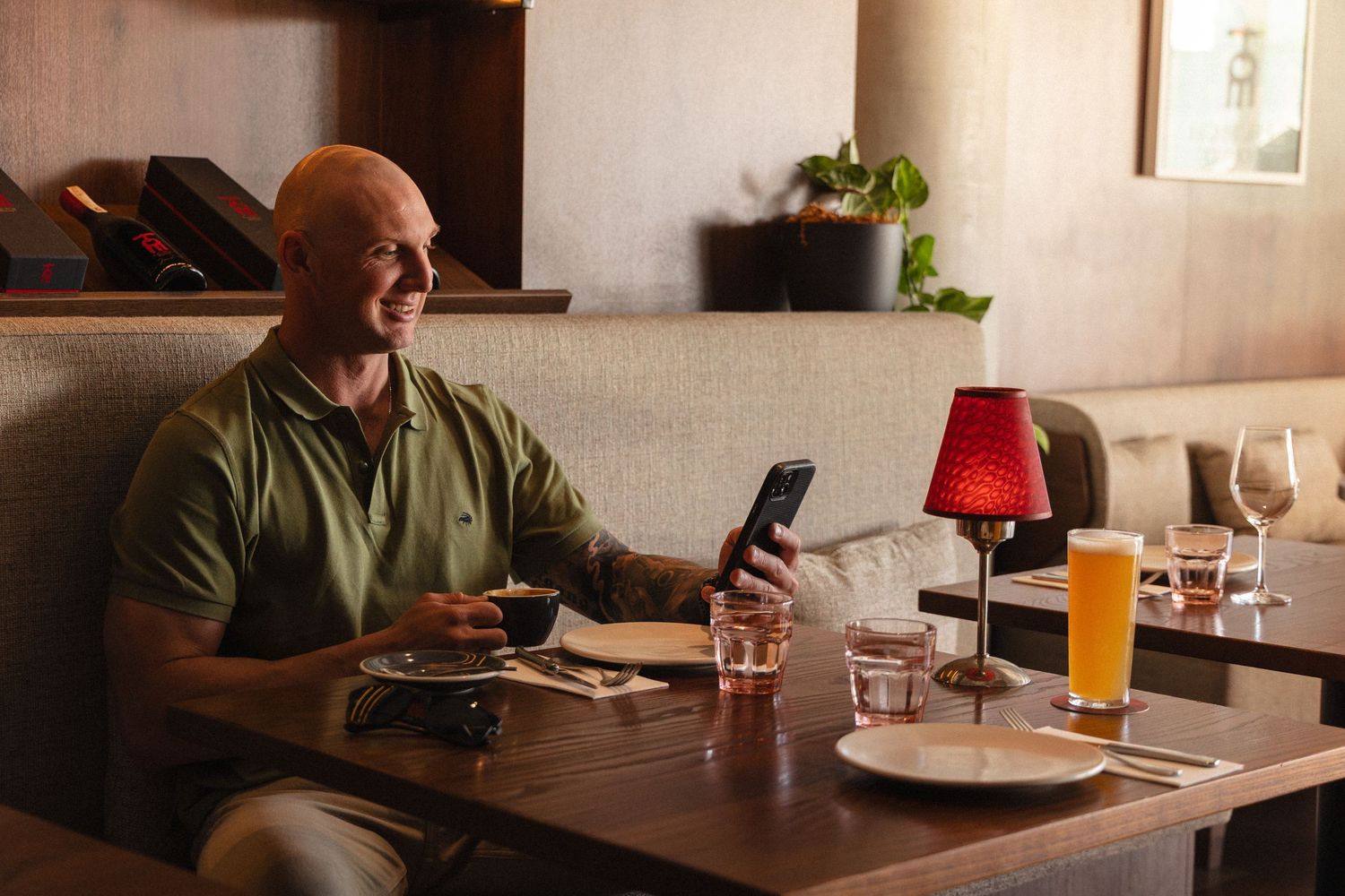 Bald man in green shirt smiling at phone while dining at restaurant table with beer.