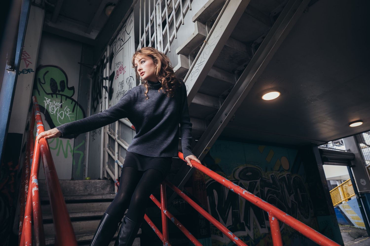 Young woman in dark outfit poses on red metal staircase in graffiti-covered urban building.
