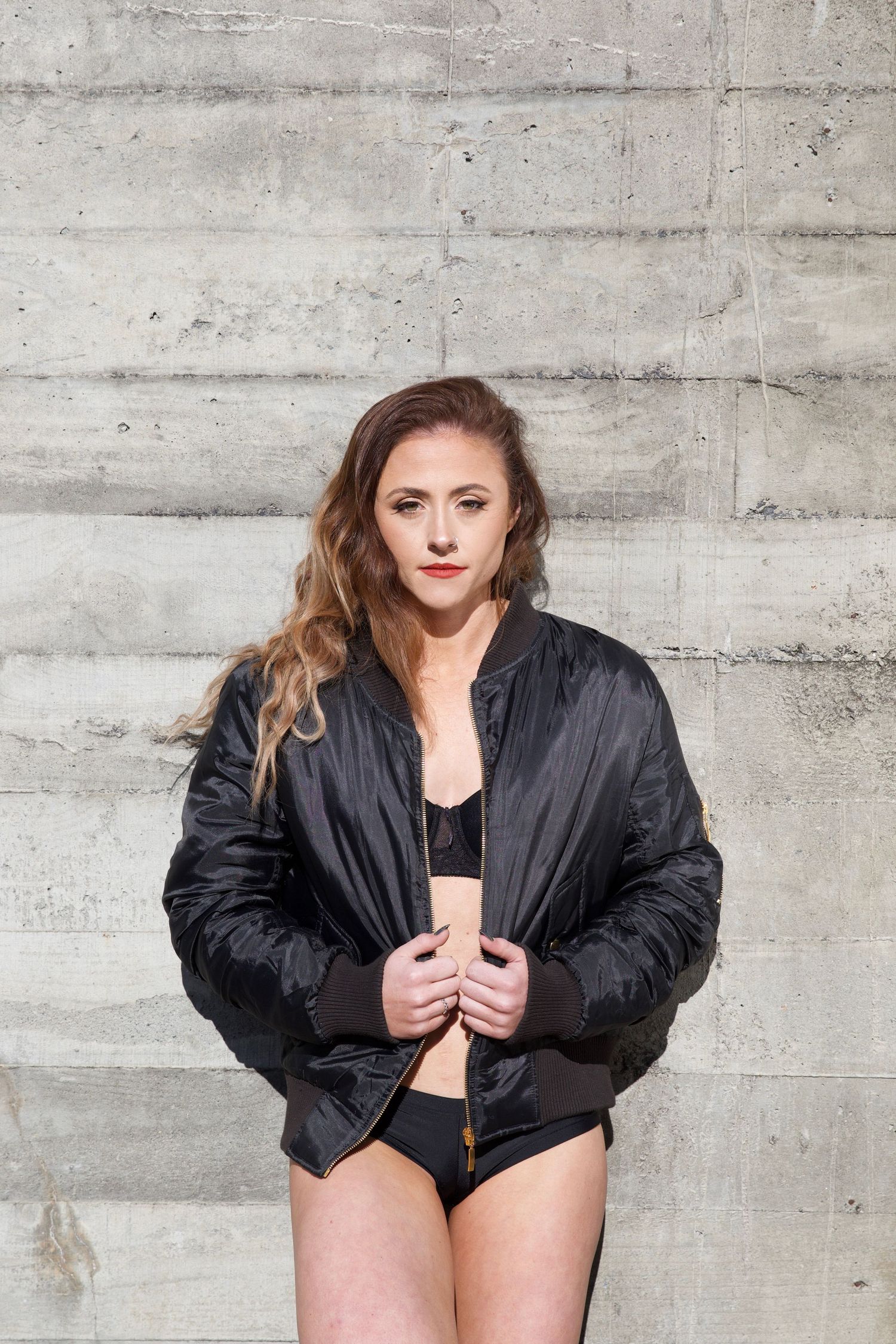 Athletic brunette woman in black bomber jacket and sports bra posing by concrete wall.