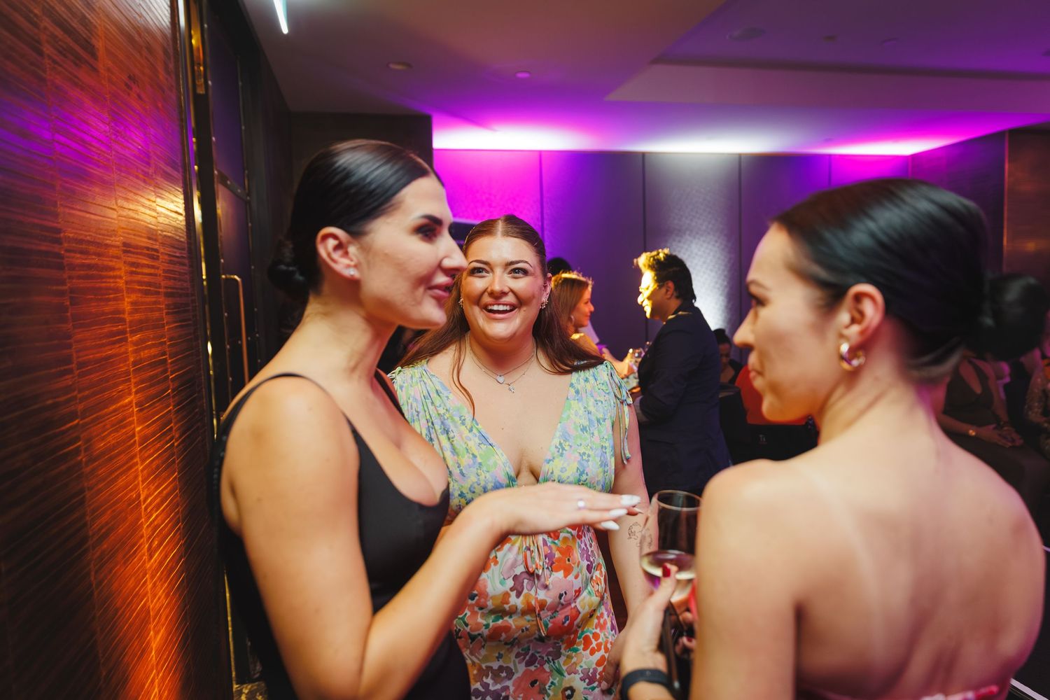 Three women engaged in conversation at a formal evening event with purple ambient lighting.