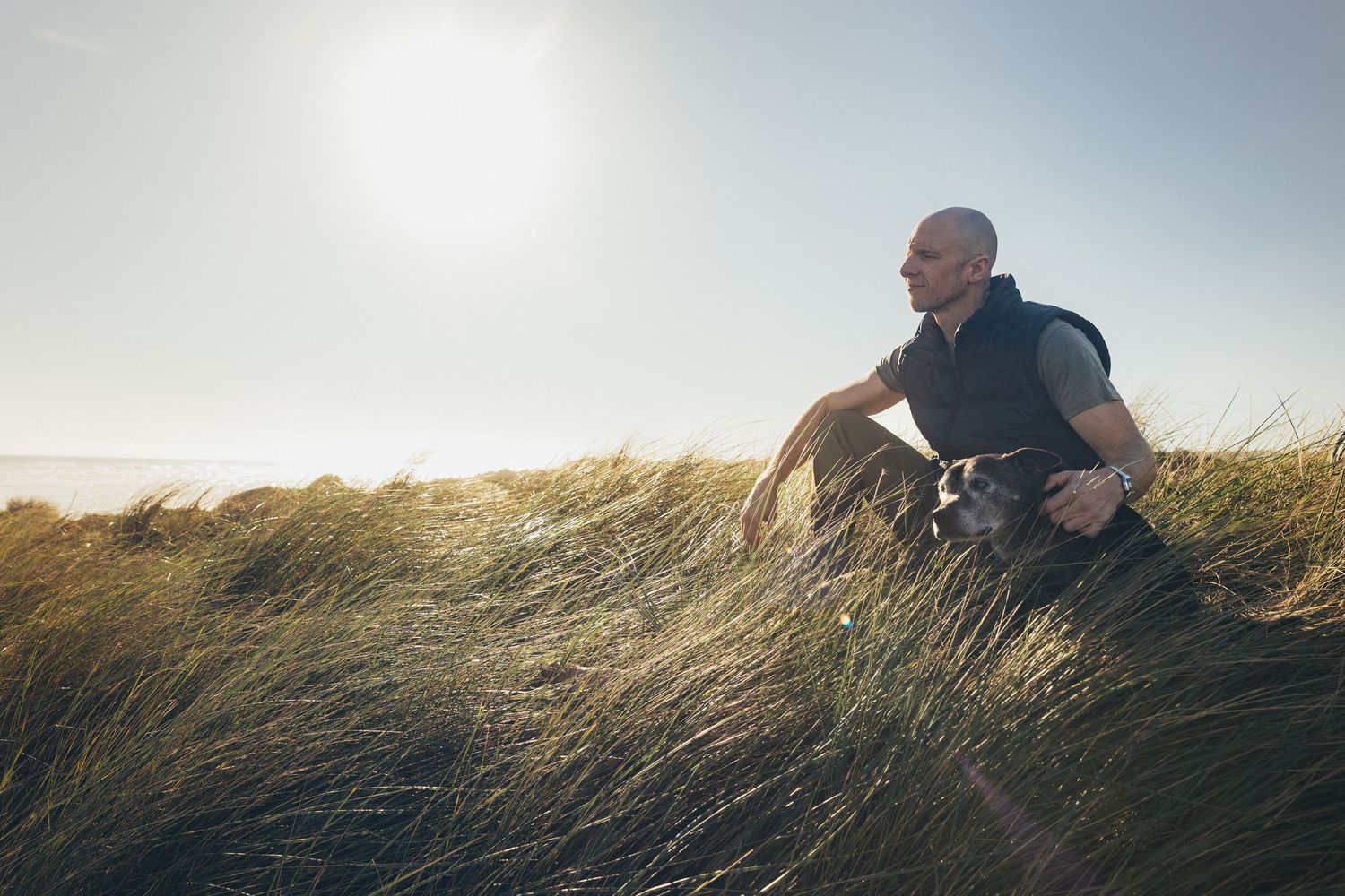 Bald man sitting with black dog in tall grass dunes on sunny coastal landscape.