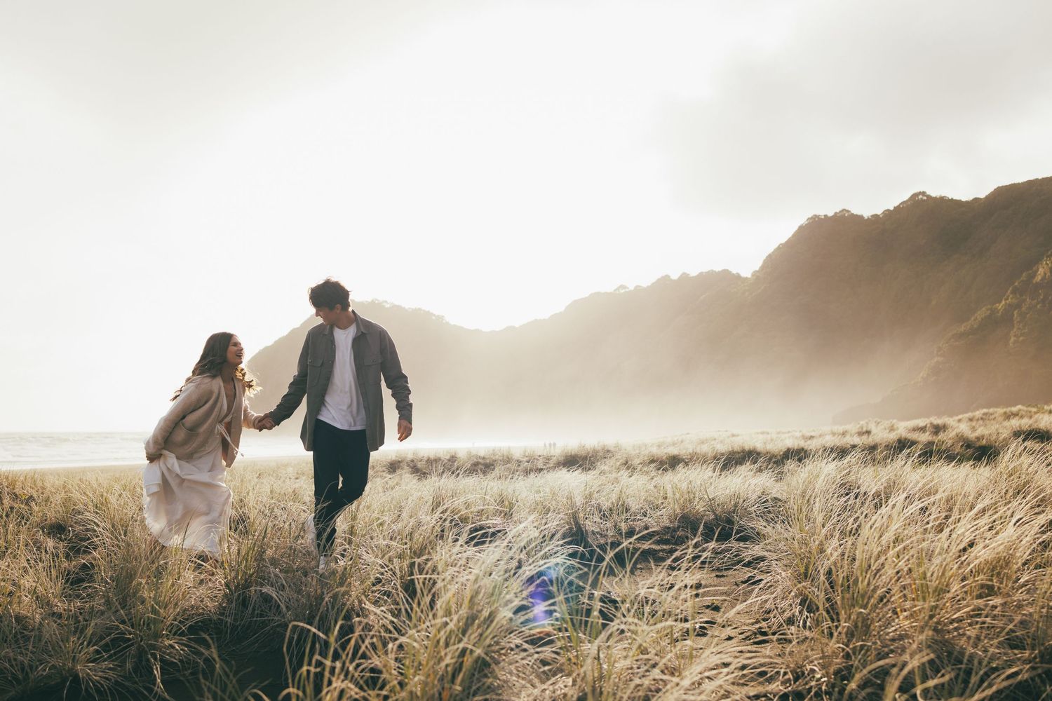 Couple holding hands walking through golden coastal grass toward misty mountains.