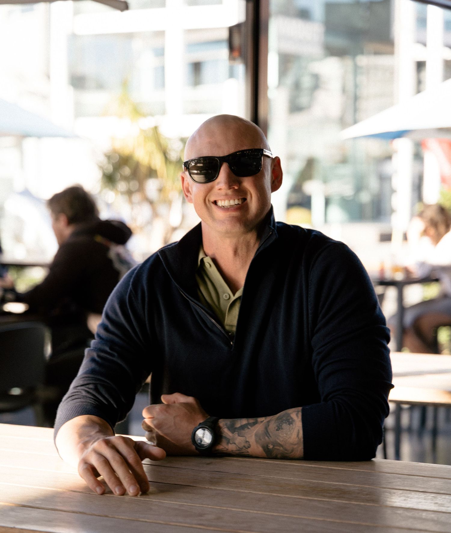 Bald man with sunglasses and tattoos smiling at outdoor cafe table, wearing navy sweater and watch.