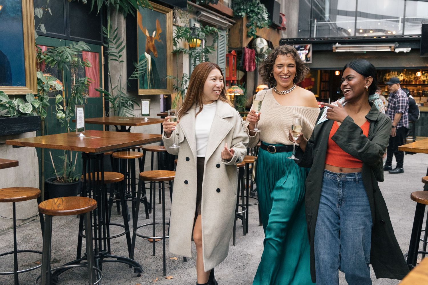 Three women laughing and strolling with champagne glasses in a trendy indoor bar setting.