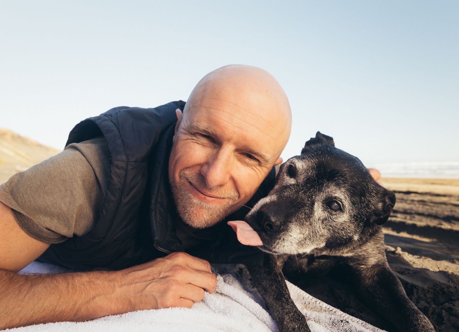 Smiling bald man lying on beach cheek-to-cheek with his senior black dog in golden light.