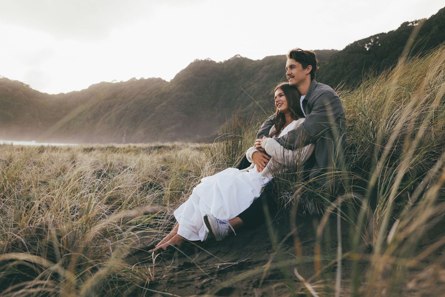 Couple sitting together in tall coastal grass with misty mountains in the background.