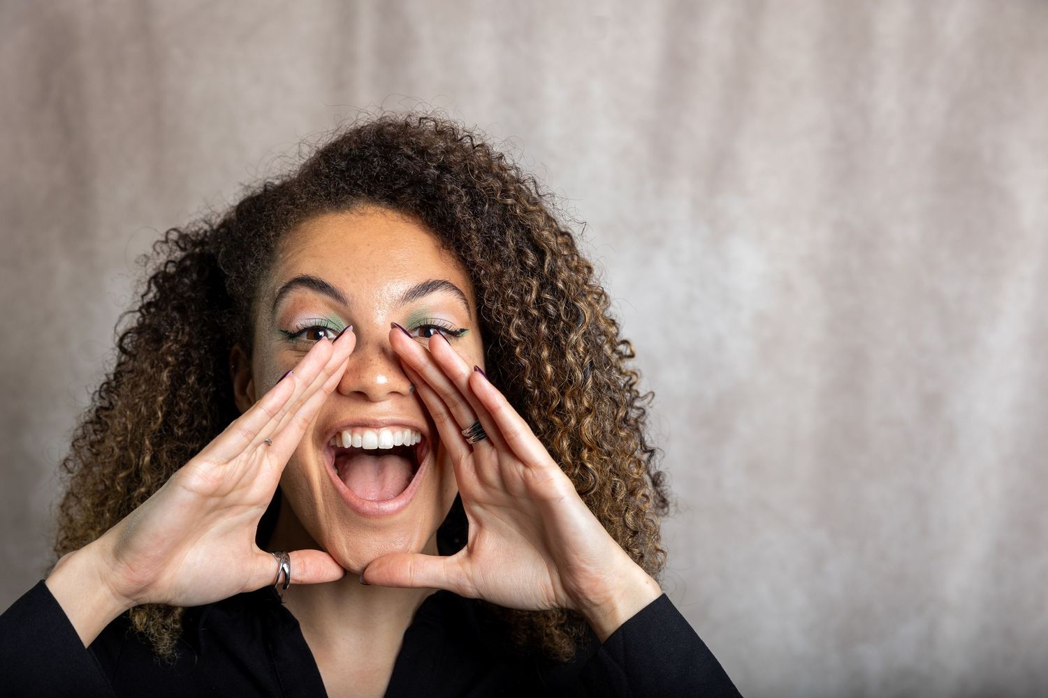 Curly-haired woman cupping hands around her mouth as if shouting against a neutral textured background.