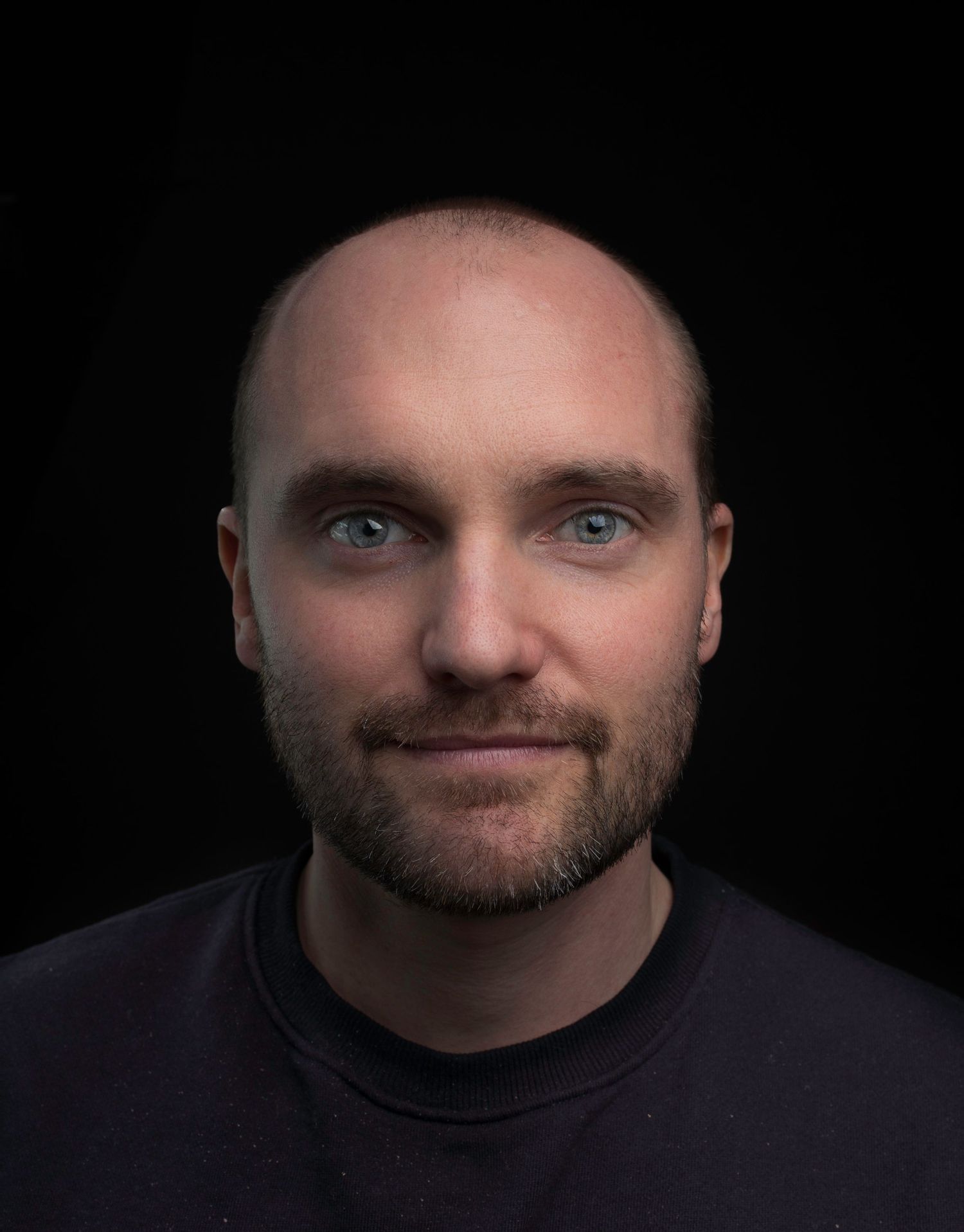 Bald man with blue eyes and short beard in black shirt against dark studio background.