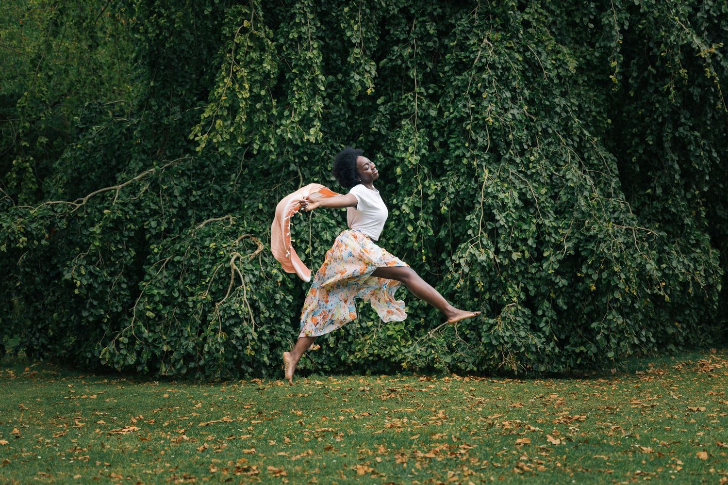 Black woman in floral dress leaping gracefully in mid-air against a backdrop of lush green weeping trees.