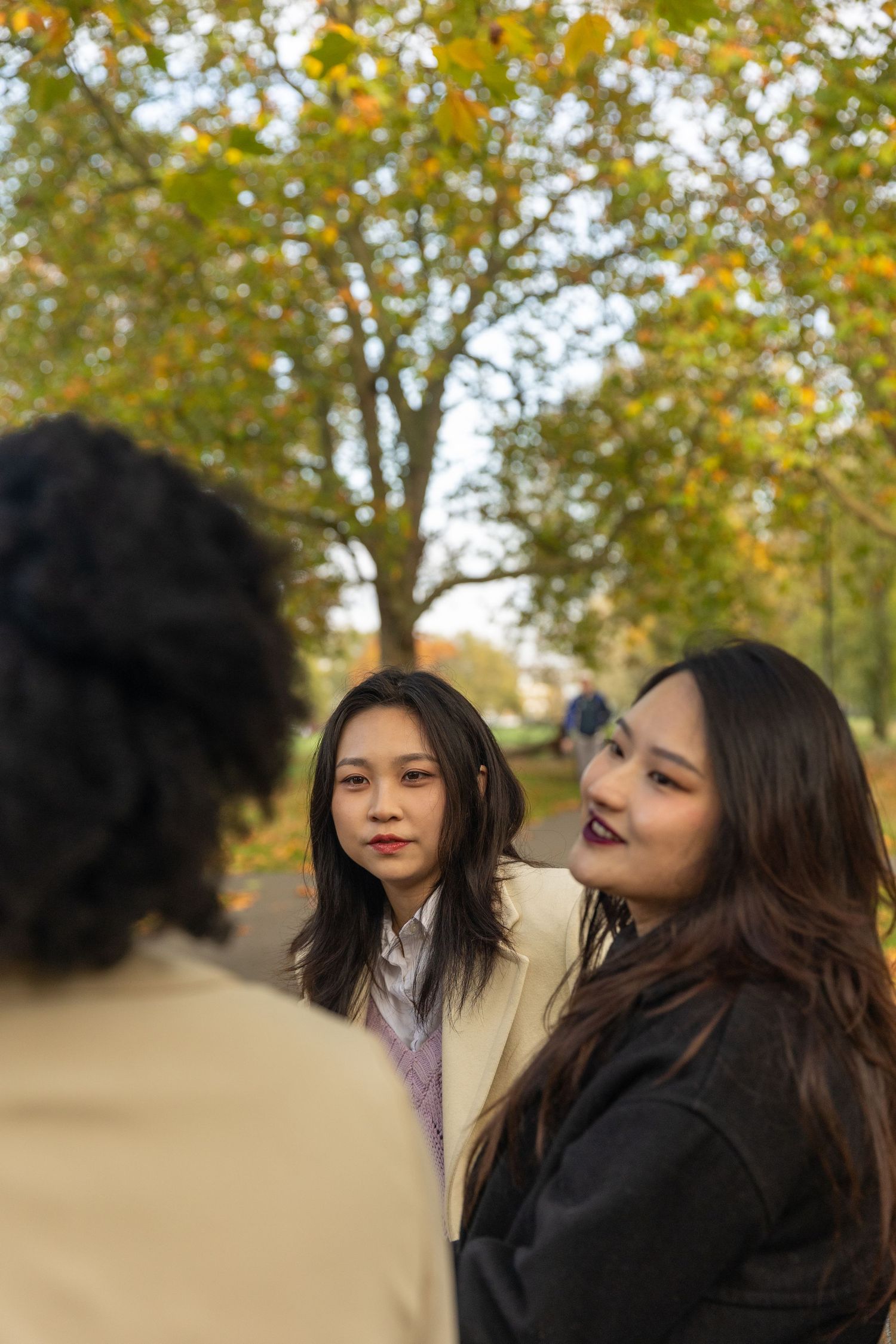 Two young Asian women in autumn coats conversing in a park with colorful fall trees behind them.