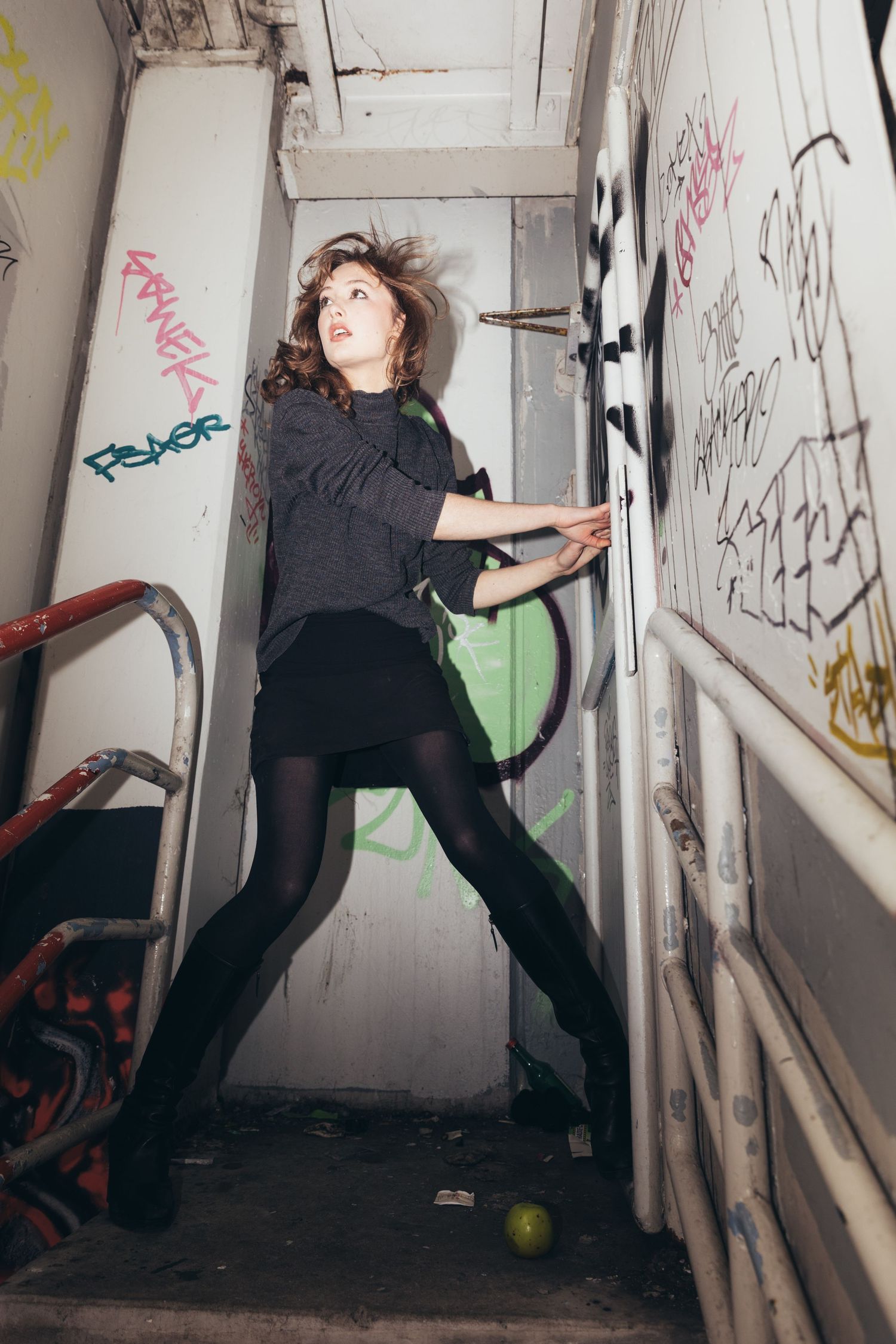 Young woman in black skirt and tights poses dramatically in graffiti-covered urban stairwell.