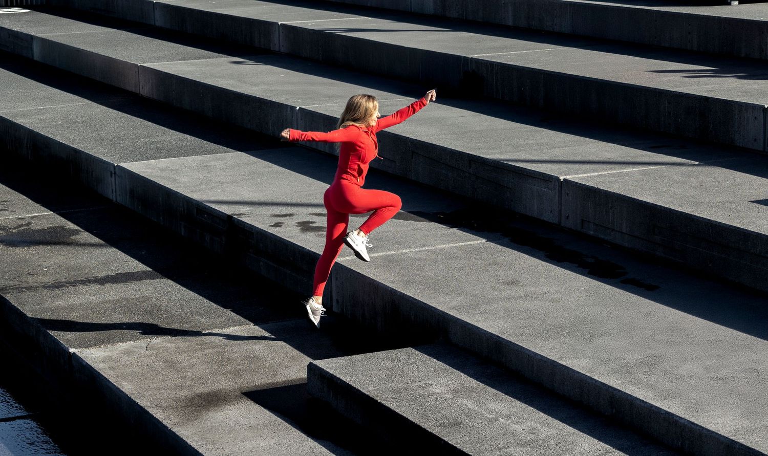 Athletic woman in red jumpsuit jumping energetically on outdoor concrete steps.