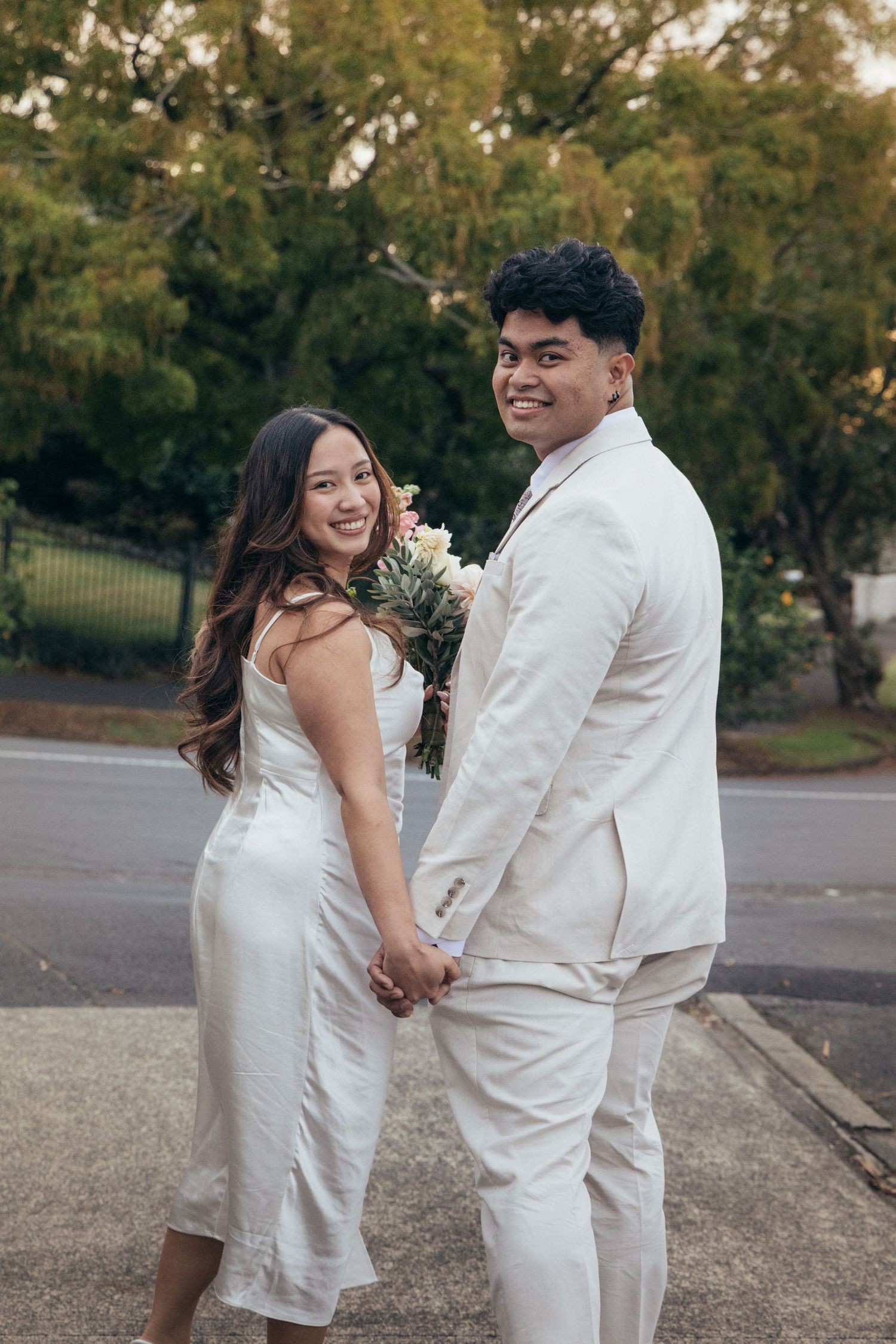 Smiling couple in white outfits holding hands outdoors, woman holding bouquet, posing for wedding or formal photo.